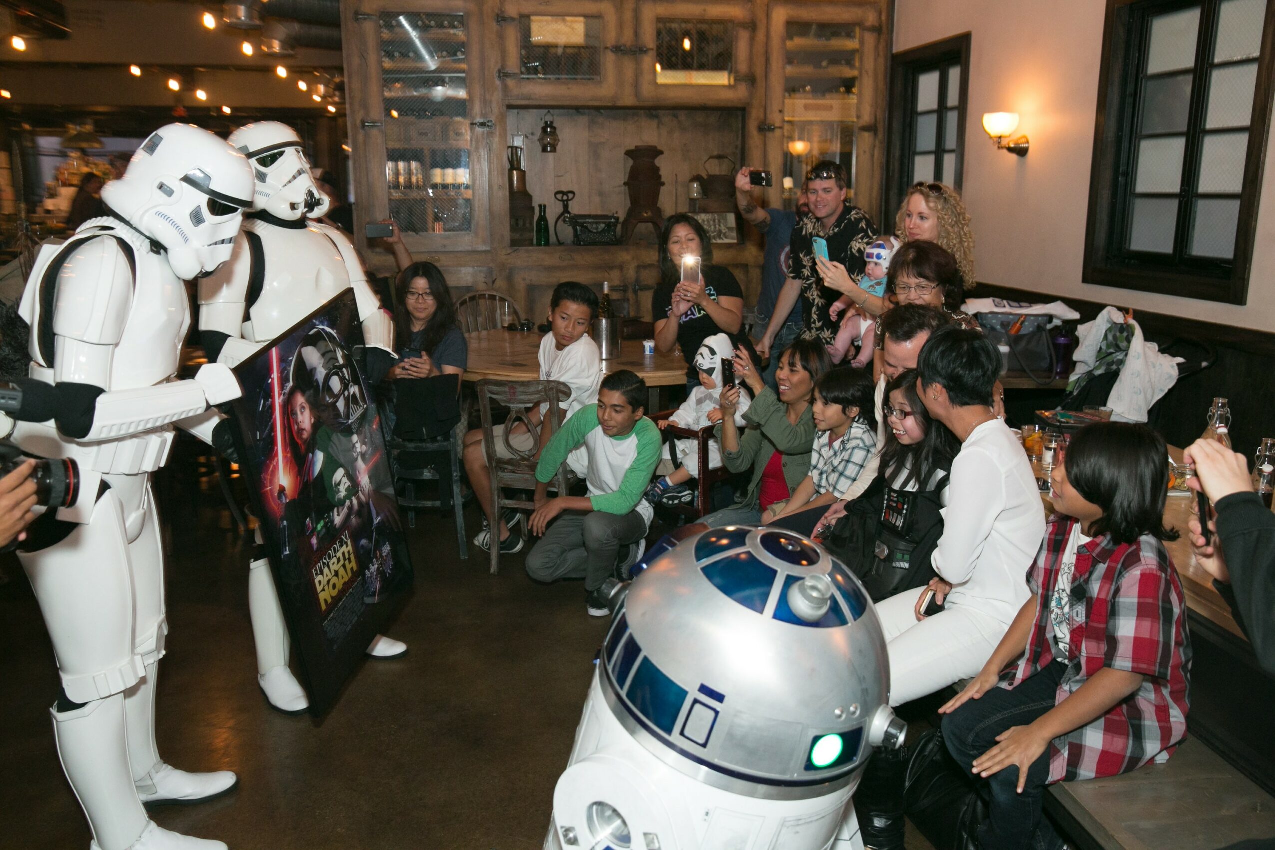 Group of children and adults watching Star Wars characters and droids in a room with wooden shelves and windows.