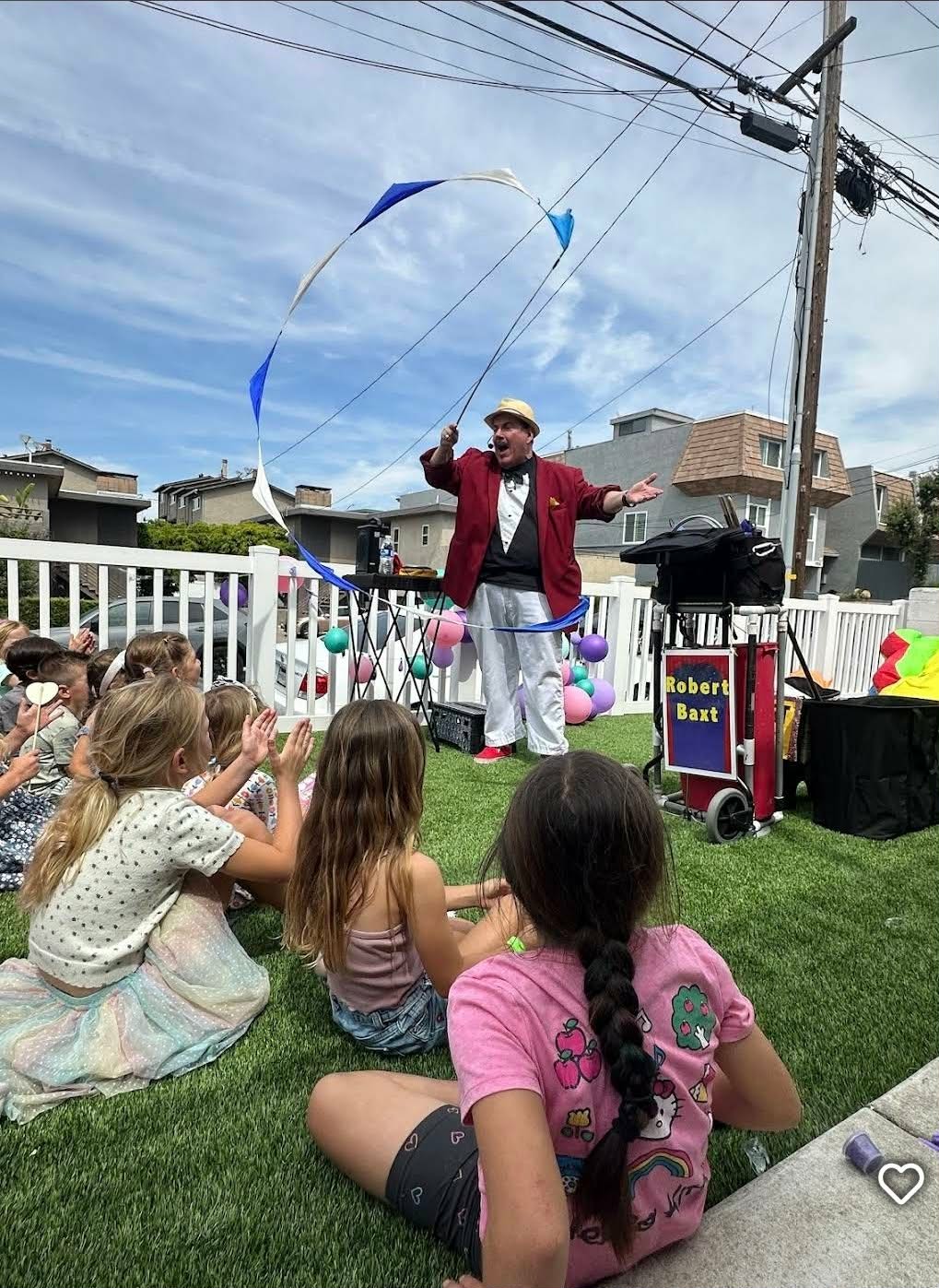 A performer in a red jacket and striped pants entertains children outdoors with a kite, in front of a white fence and houses.