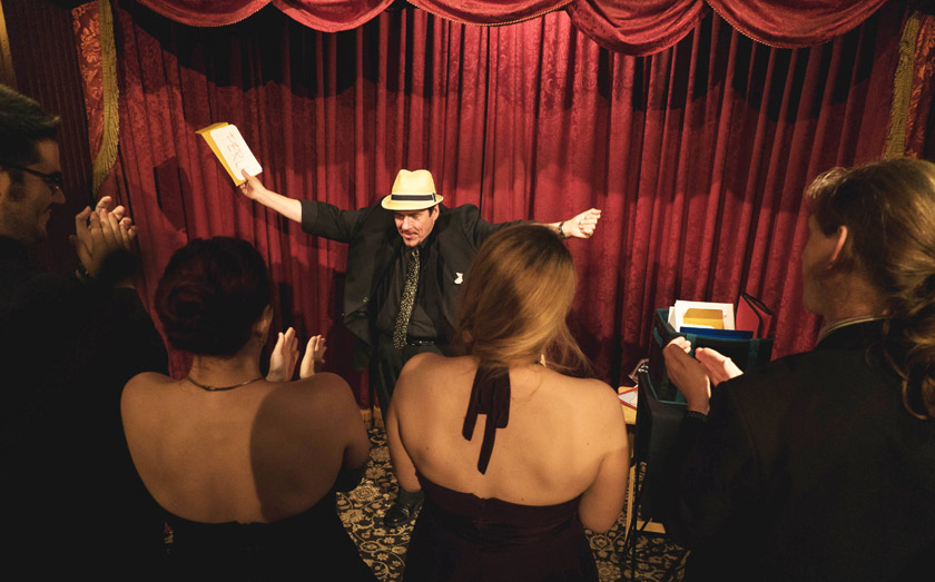 Performer on stage with arms outstretched, holding a paper, in front of a red curtain, surrounded by audience members.