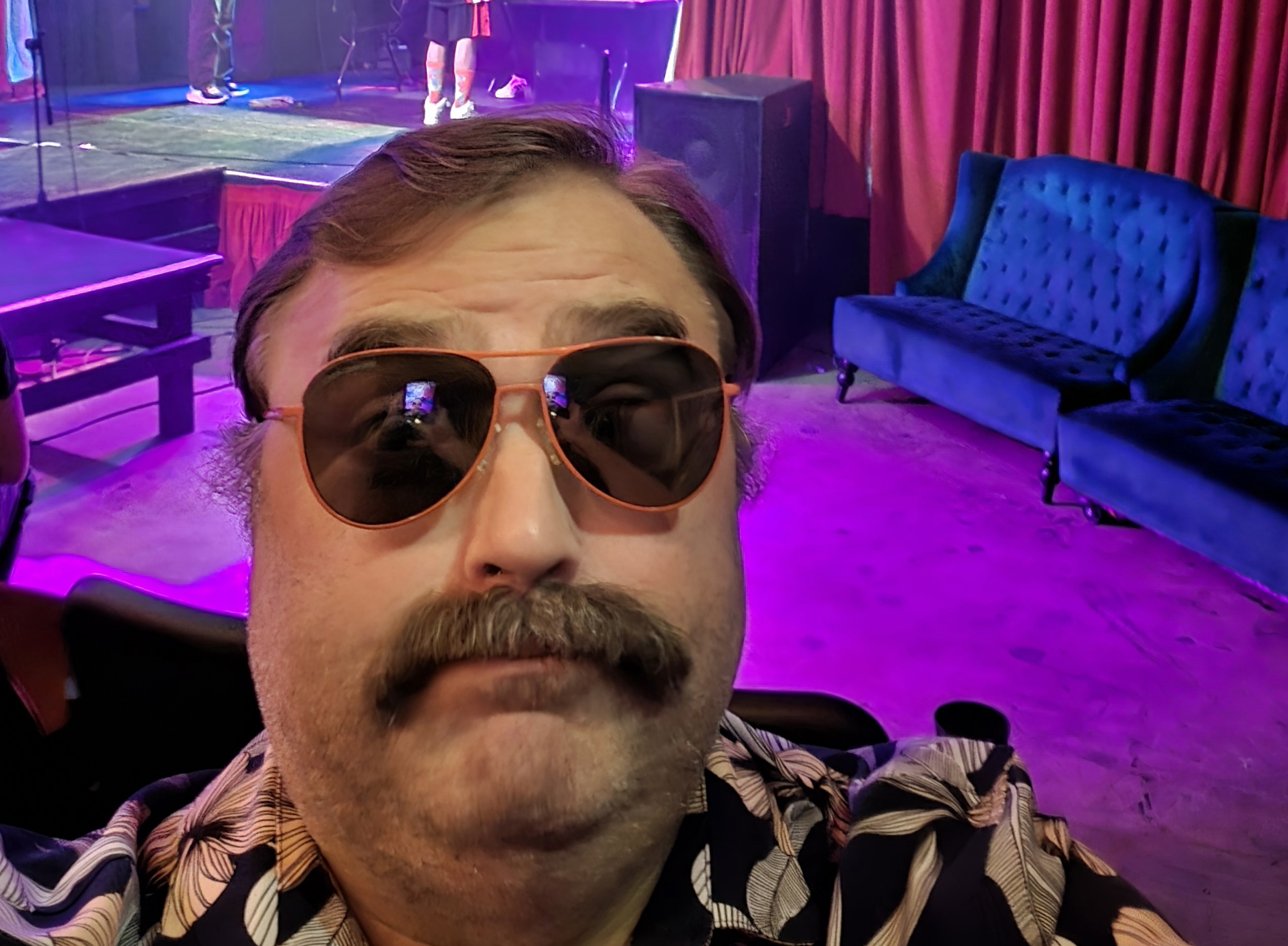 Person with sunglasses and mustache taking a selfie in a colorful, dimly lit venue with couches and curtains.