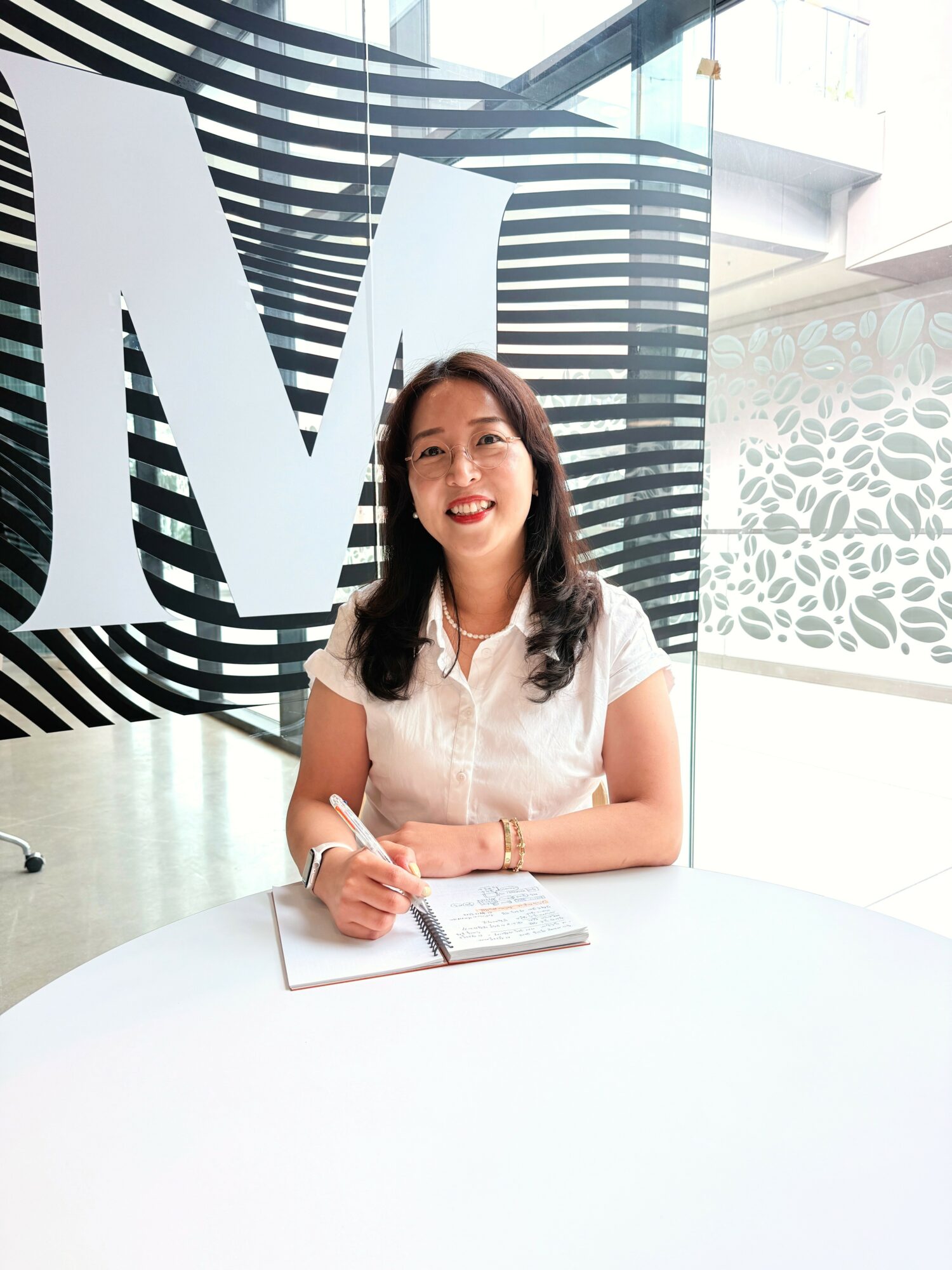 Woman sitting at a table with a notebook, smiling, in a modern indoor space with large black and white patterned background.