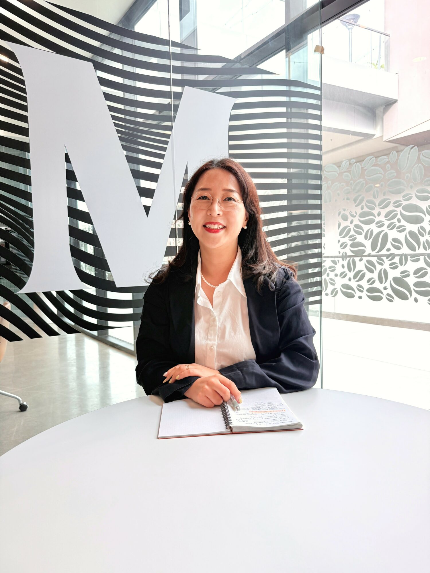 Woman sitting at a table with a notebook, smiling, in a modern office with large windows and black-and-white wall art.