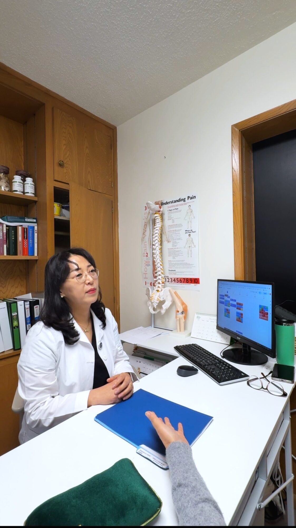 Woman sitting at desk with computer, bookshelf, and spine model in room with white walls and wooden door.