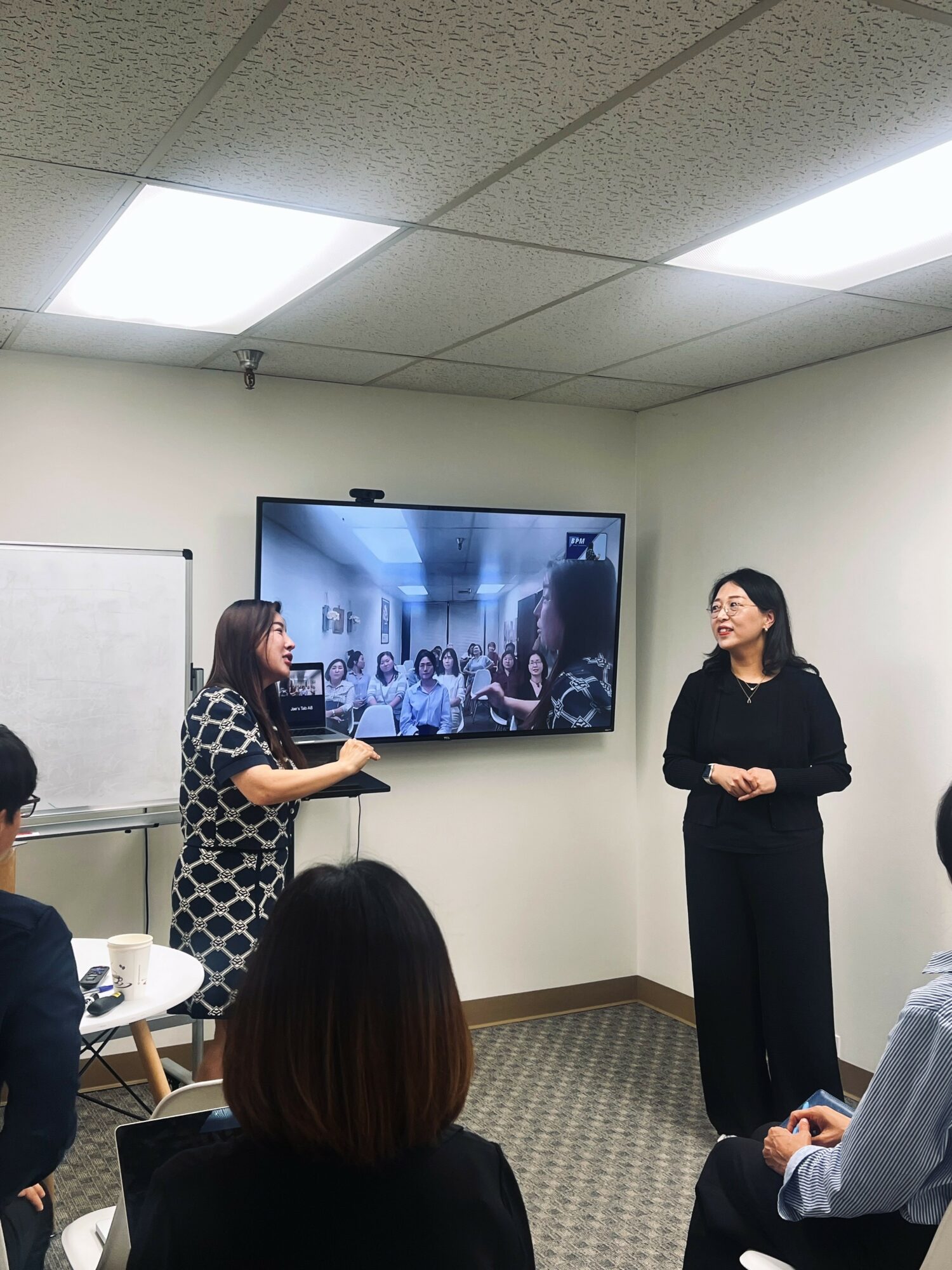 Two women standing and speaking in a room with a large screen and audience, white walls, and ceiling lights.
