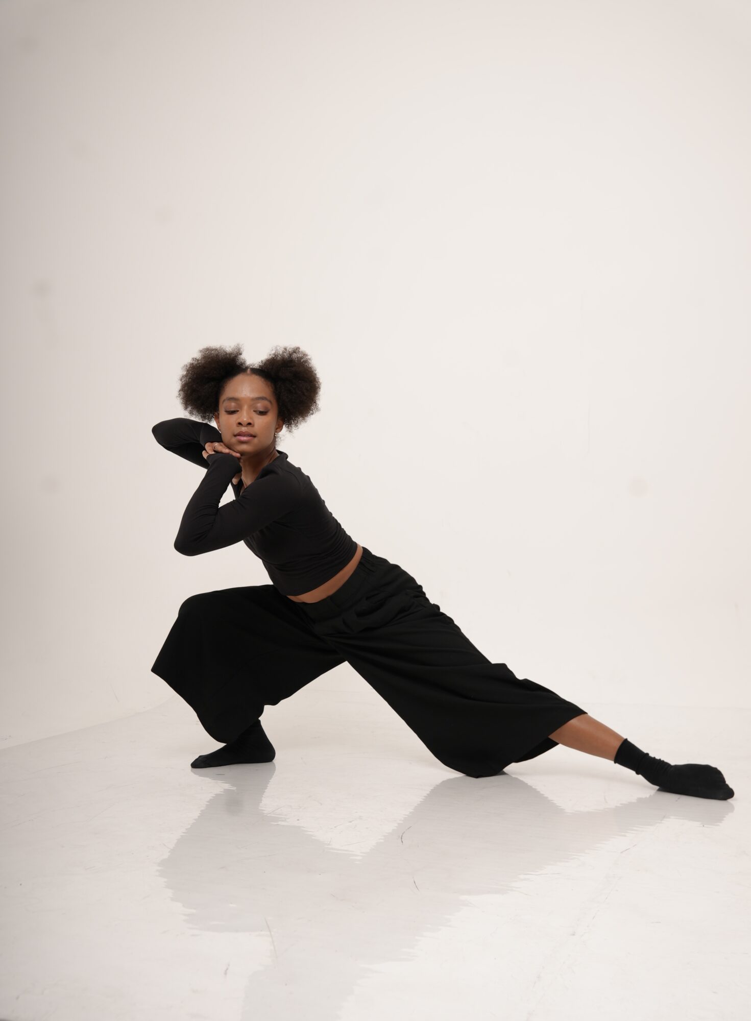 Young woman in black dance outfit poses on white background, one arm bent behind head, one extended, wide stance, looking at camera.