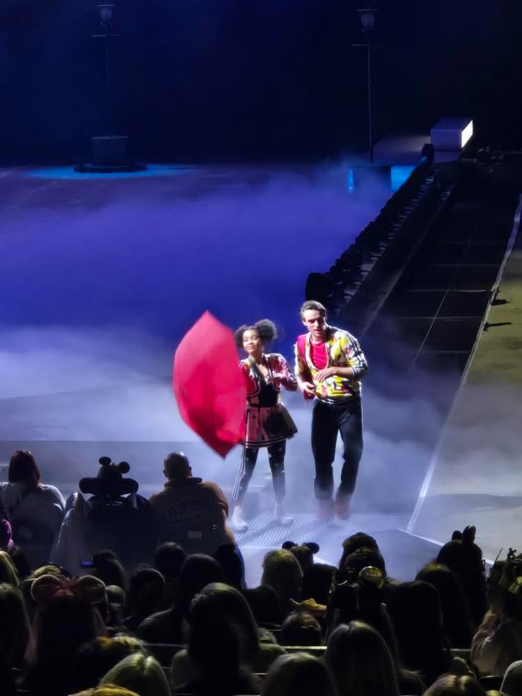 Two performers on stage, one holding a red umbrella, with an audience watching in front, stage fog and stairs in background.