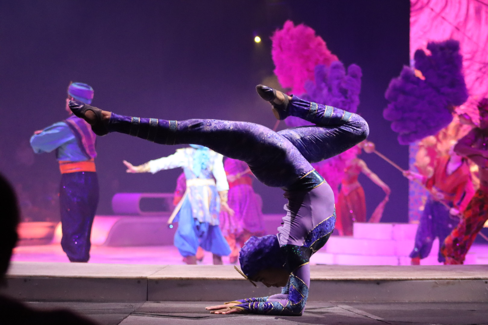 Person performing a headstand on stage with colorful background and other performers in the background.