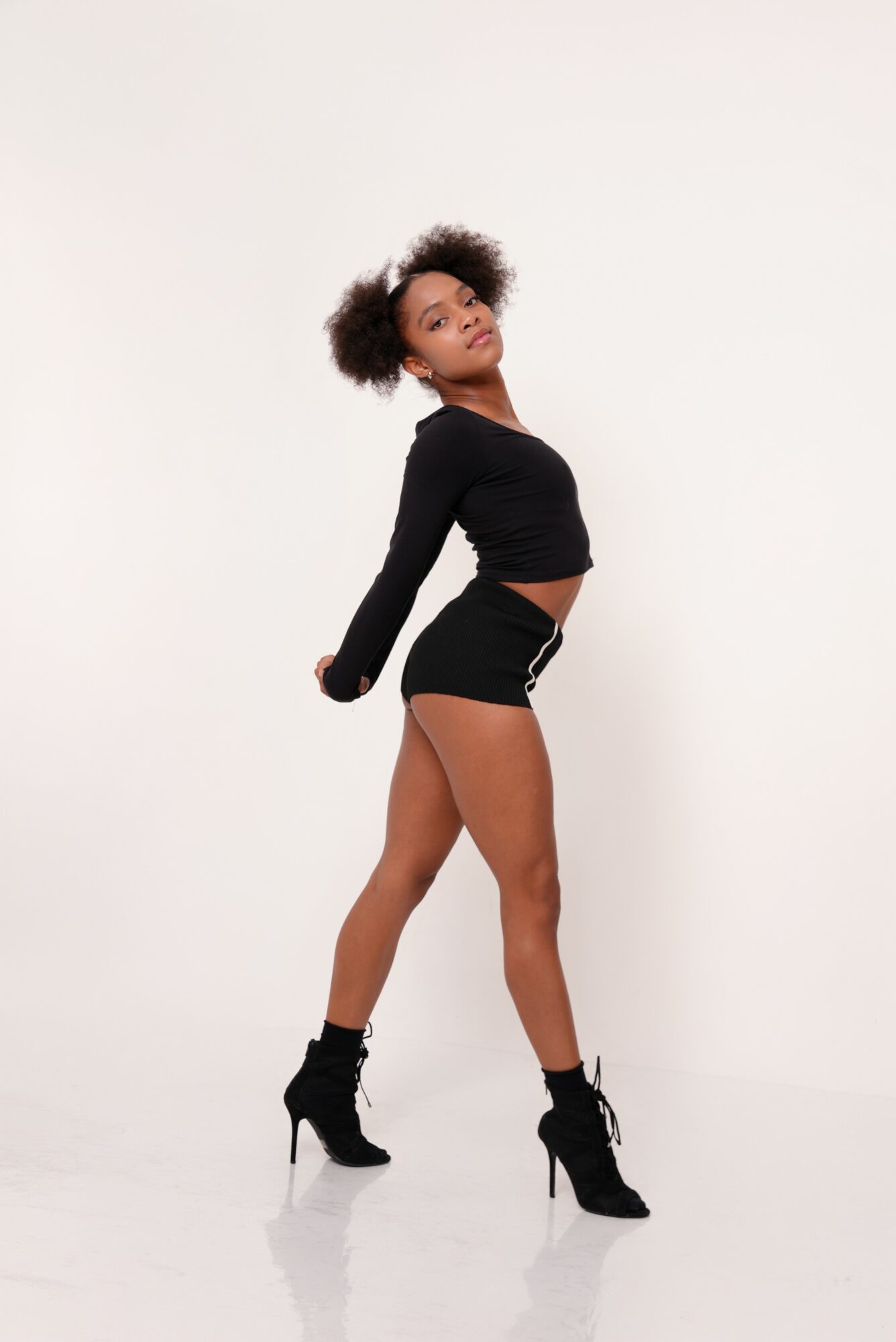 Young woman with natural hair in a black long-sleeve crop top and shorts, standing sideways with one leg forward, wearing high heels.