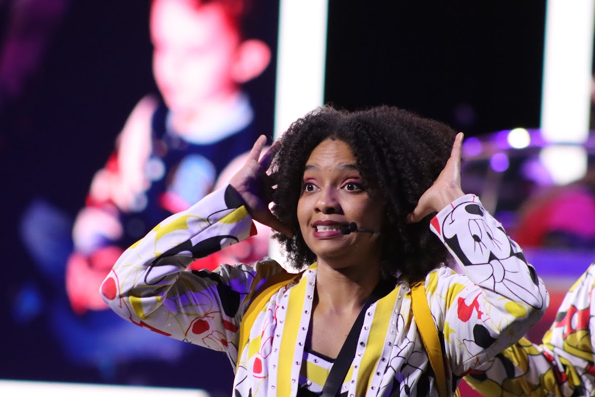 Woman with curly hair smiling and holding her hands near her head, wearing a colorful patterned jacket, on stage with a blurred background.