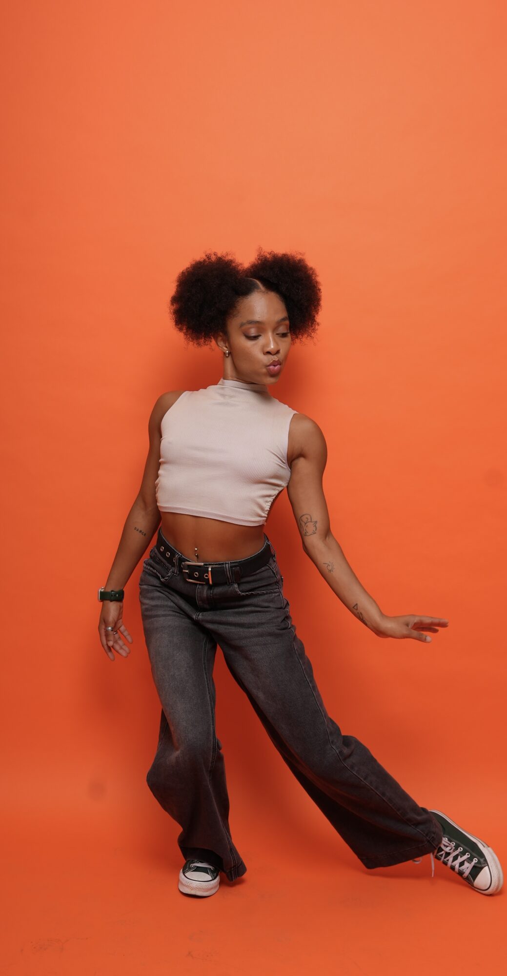 Young woman with curly hair in a sleeveless top and jeans poses against an orange background.