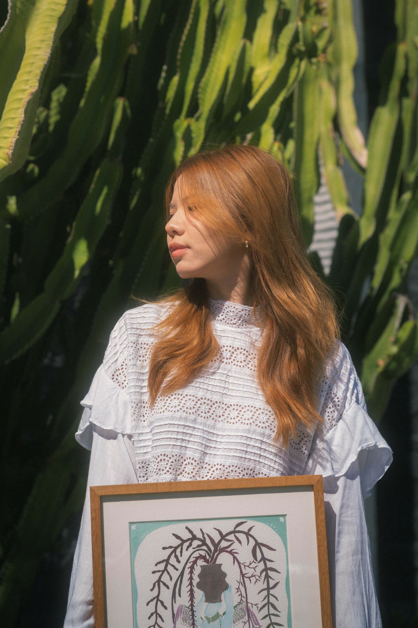 Woman with long hair standing outdoors in front of tall green plants, holding a framed artwork.