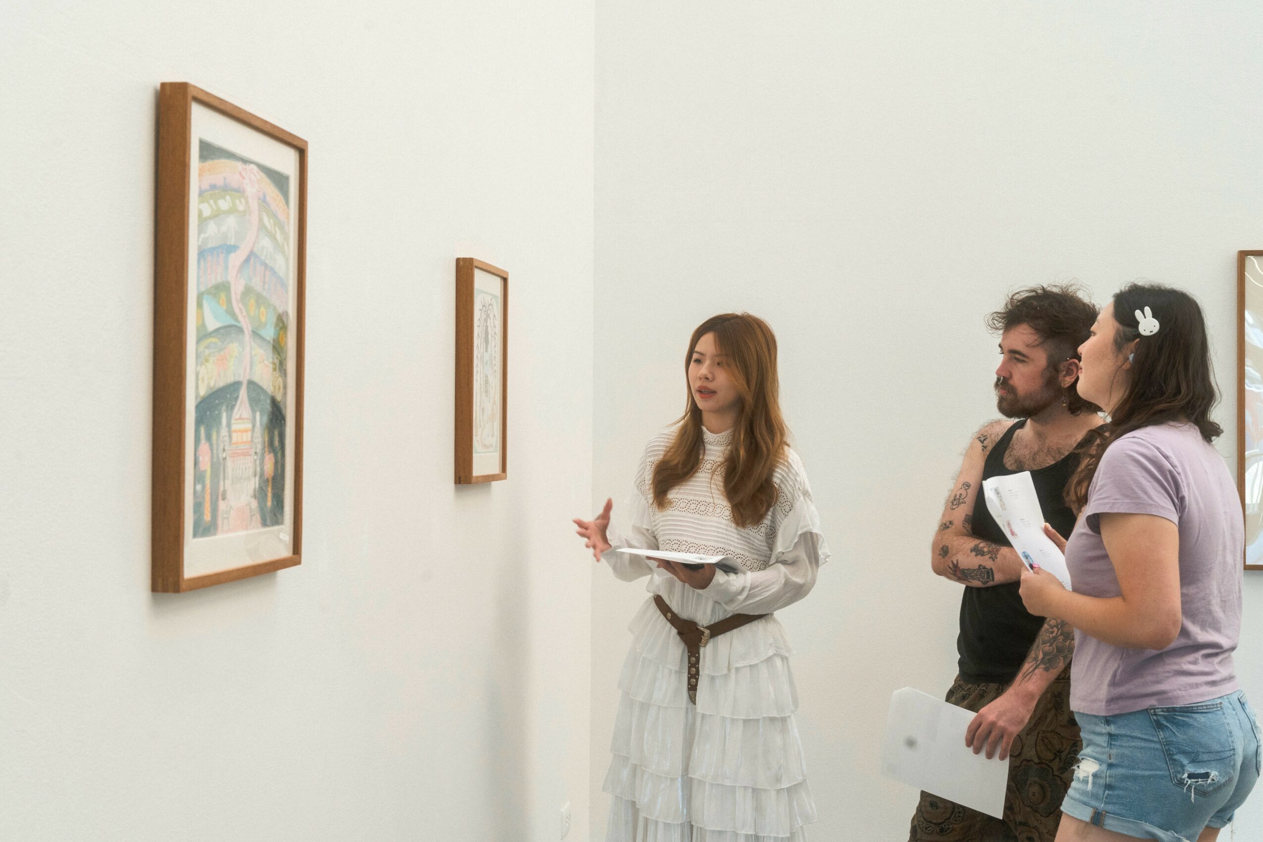 Three people in an art gallery looking at paintings on the wall, one woman explaining, two listening attentively.