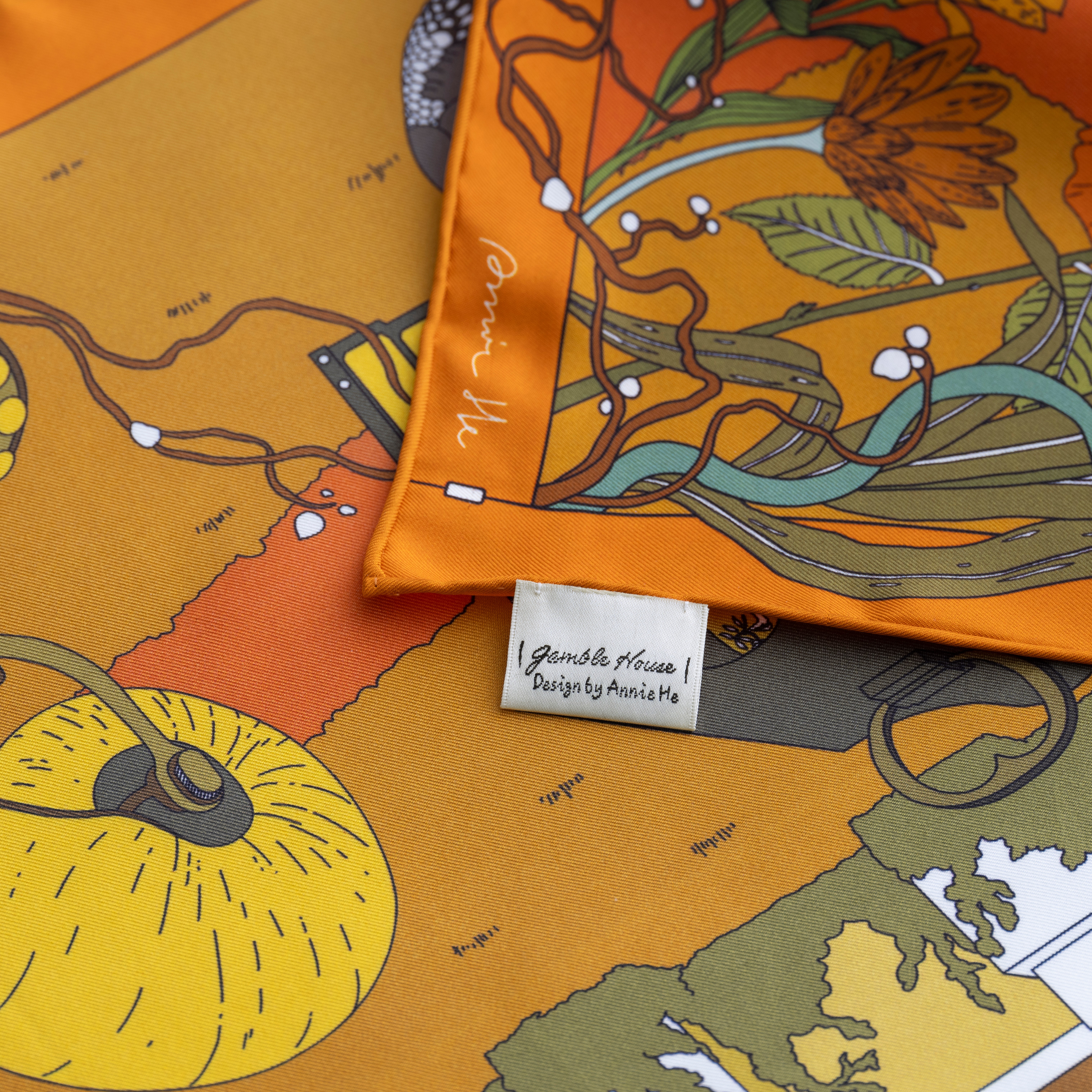 Close-up of a colorful fabric with floral and fruit patterns, including a yellow fruit and orange flowers, and a white tag.