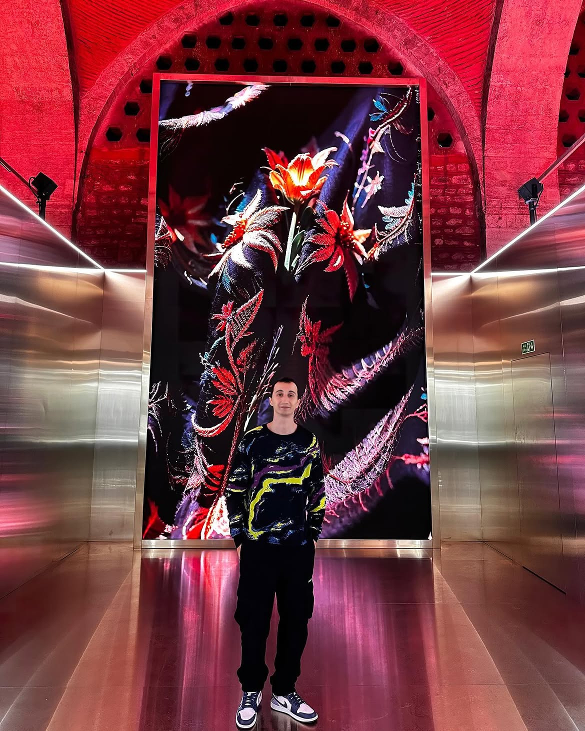 Person standing in front of a large, colorful digital display with floral and abstract designs, inside a red brick archway.