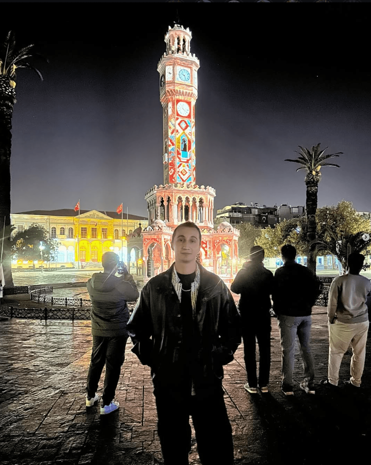 Person standing in front of a colorful clock tower at night with palm trees and buildings in the background.