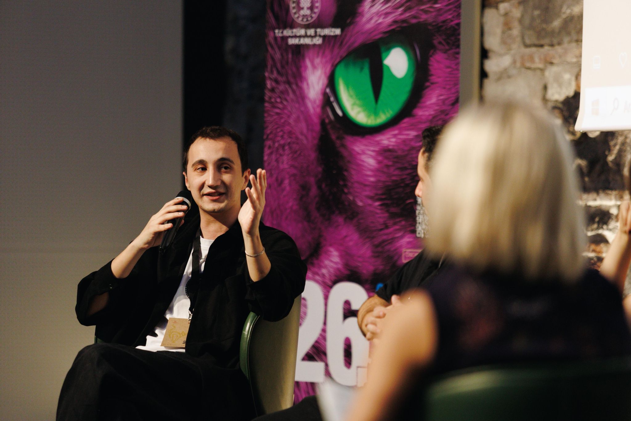 Person speaking with a headset, smiling, and gesturing, with a woman in the foreground and a large colorful poster behind.