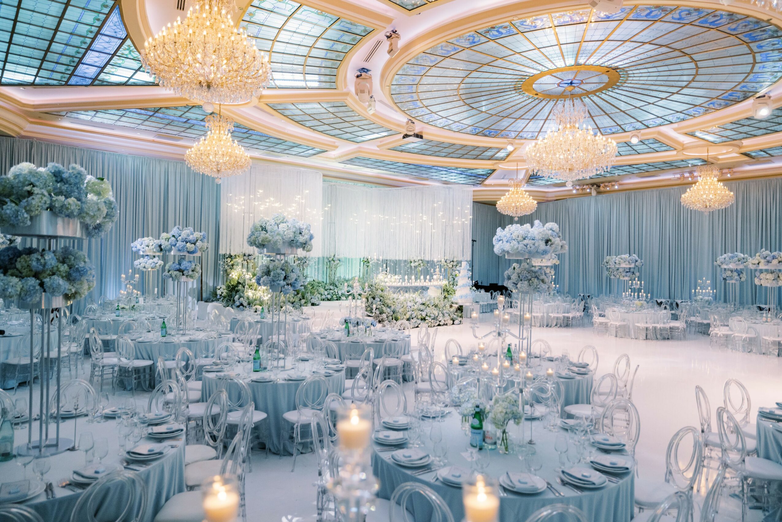 Elegant banquet hall with chandeliers, round tables, floral centerpieces, and soft lighting, decorated for a formal event.