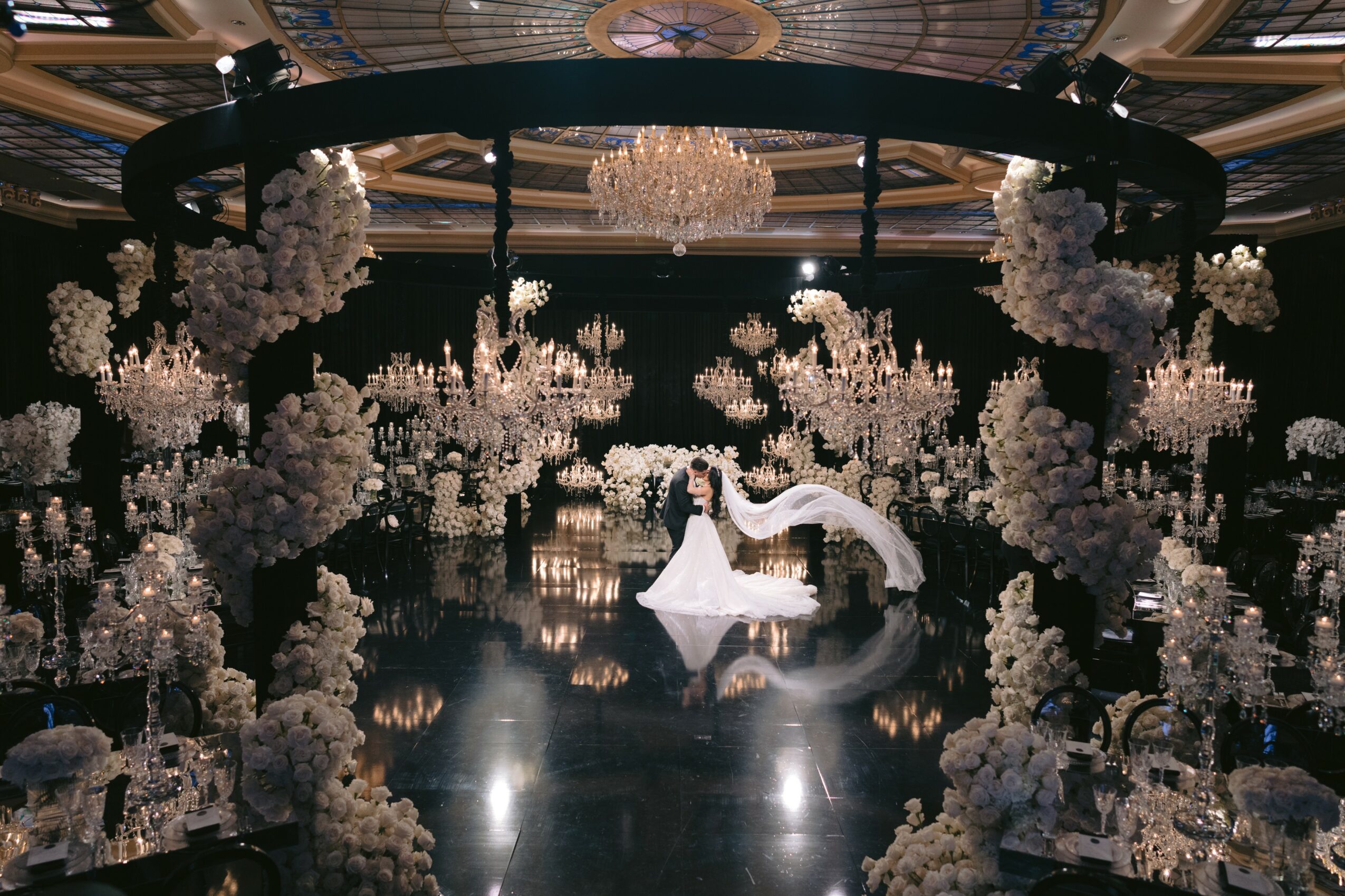 A bride and groom kiss under a circular floral arch with chandeliers in an elegant ballroom.