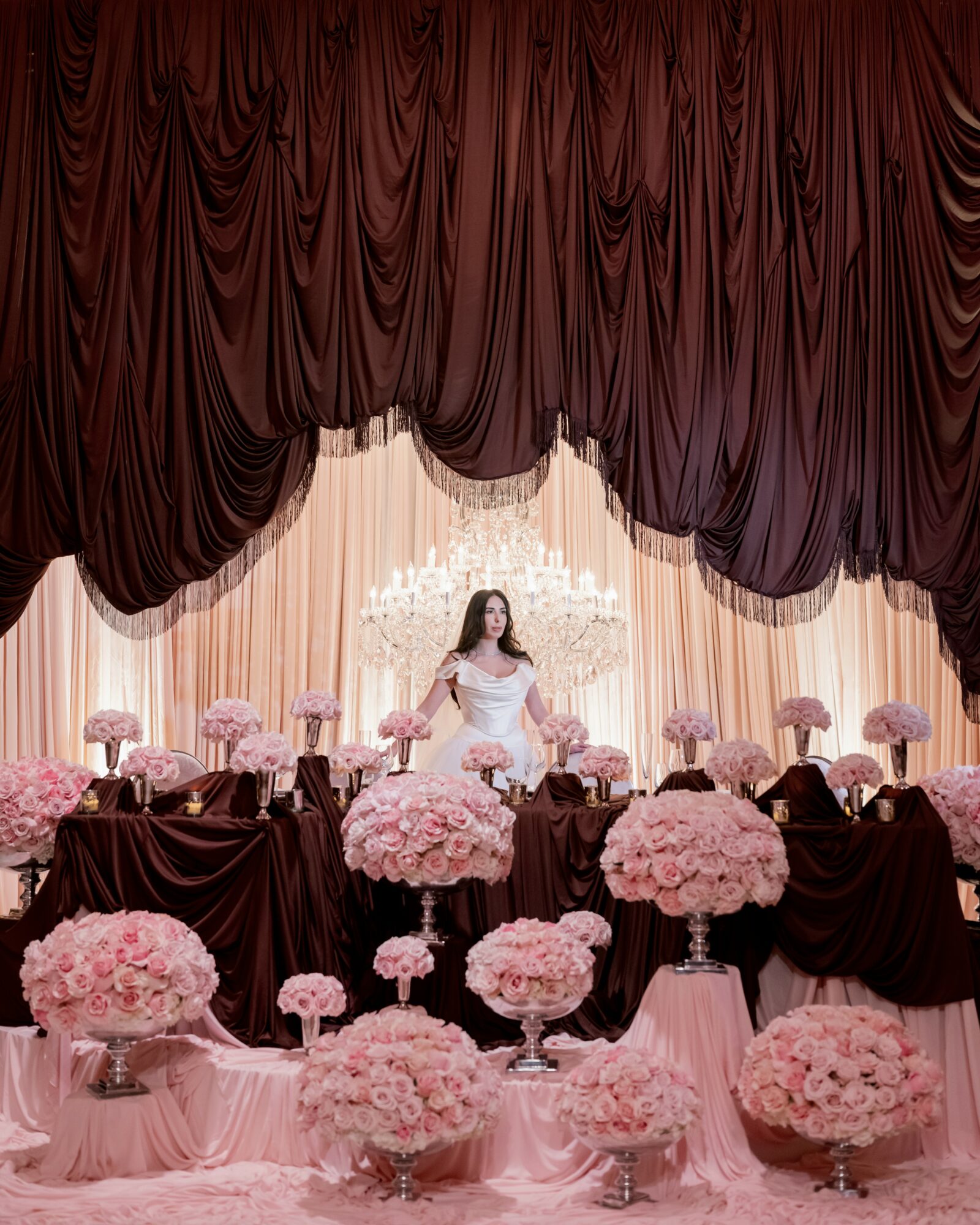 A woman in a white dress stands behind a table decorated with pink flowers, in front of a red curtain backdrop.