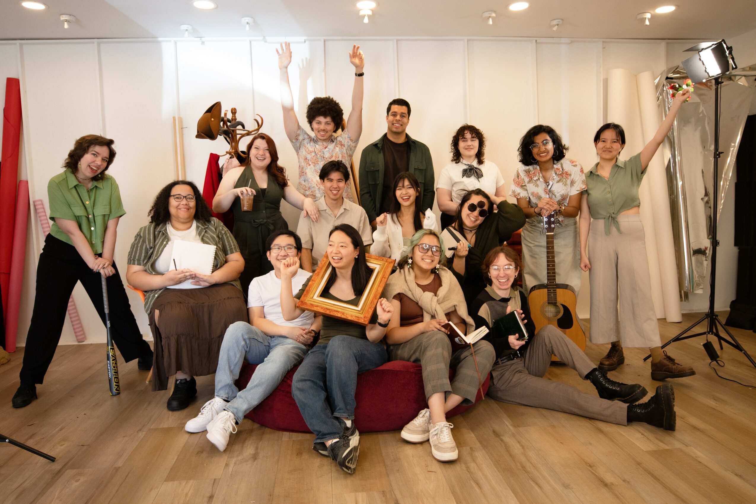 Group of 14 people posing indoors, some holding awards or certificates, some making gestures, with a white background and wooden floor.