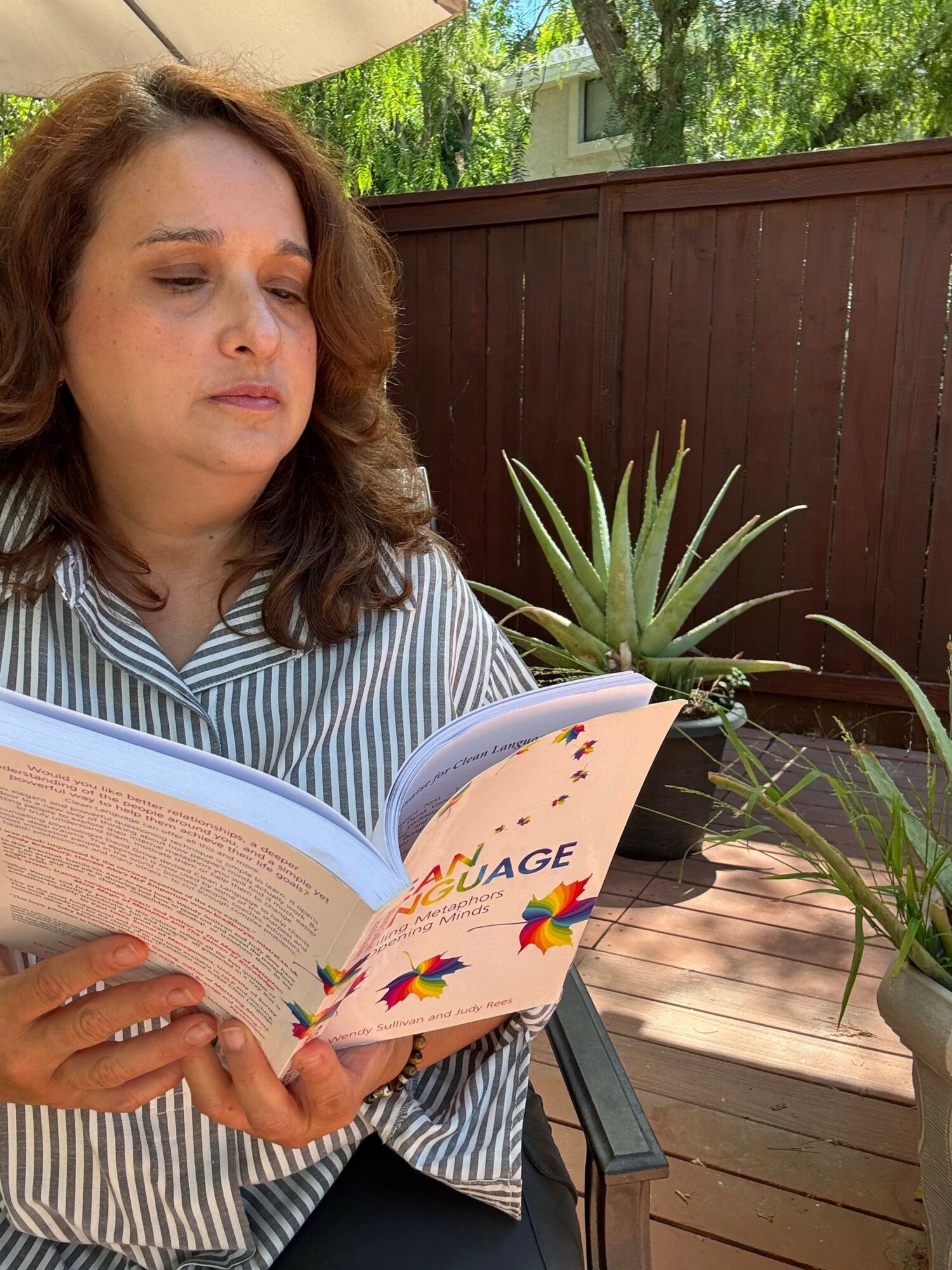 Woman with brown hair reading a colorful book outdoors on a wooden deck, with plants and trees in the background.