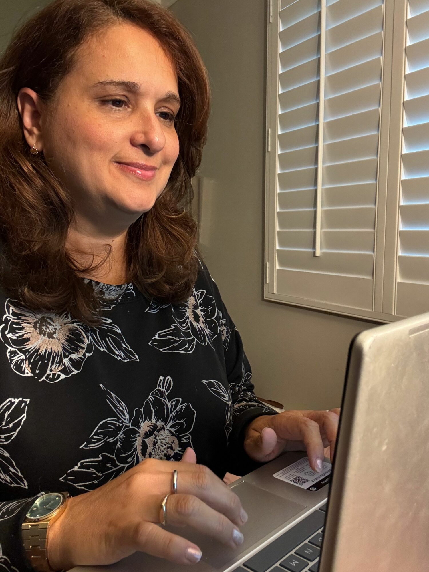 Woman with brown hair taking a selfie, sitting at a desk with a laptop, window blinds in background.