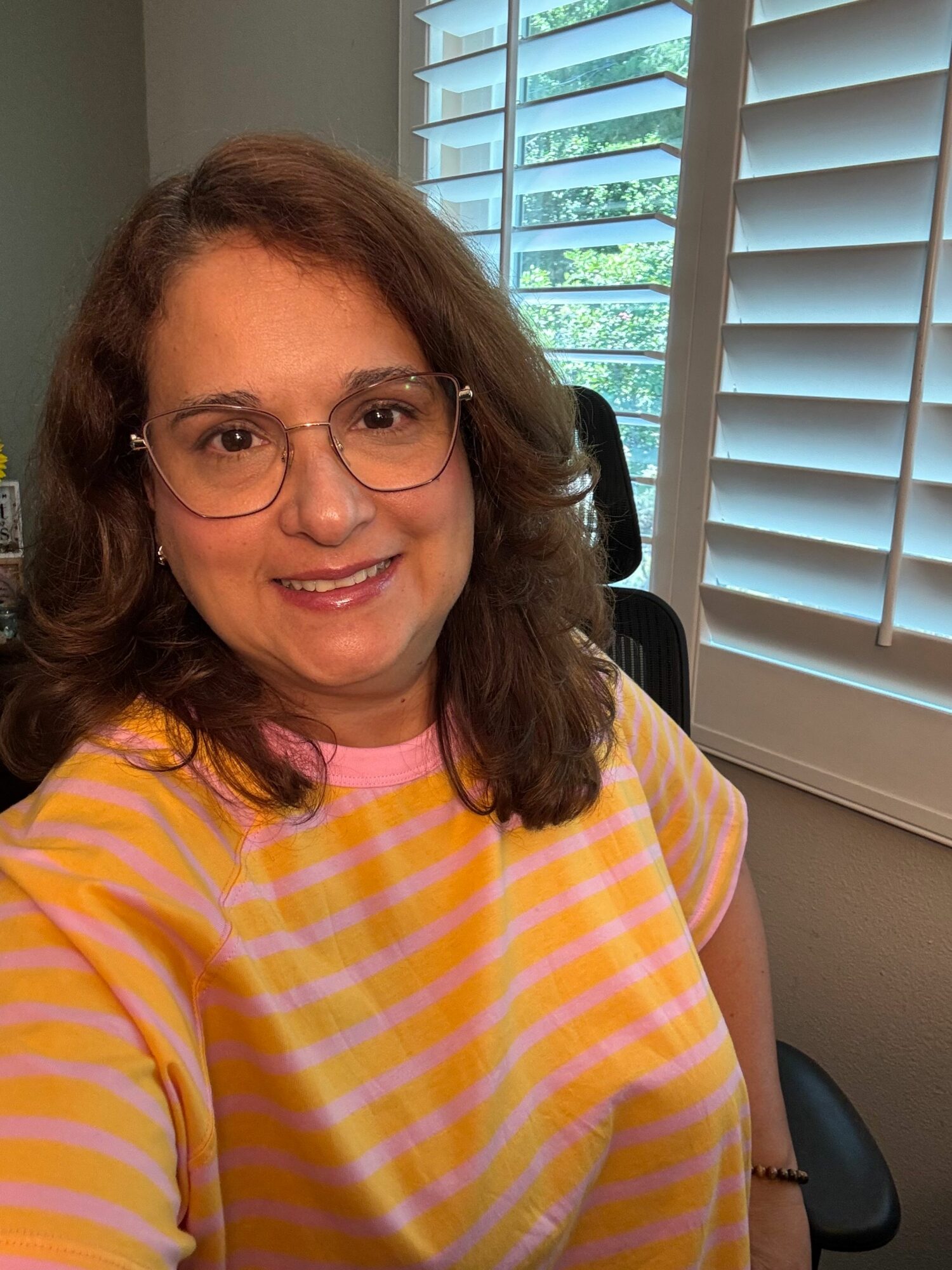 Woman with glasses smiling, wearing a yellow and pink striped shirt, sitting indoors near a window with blinds.