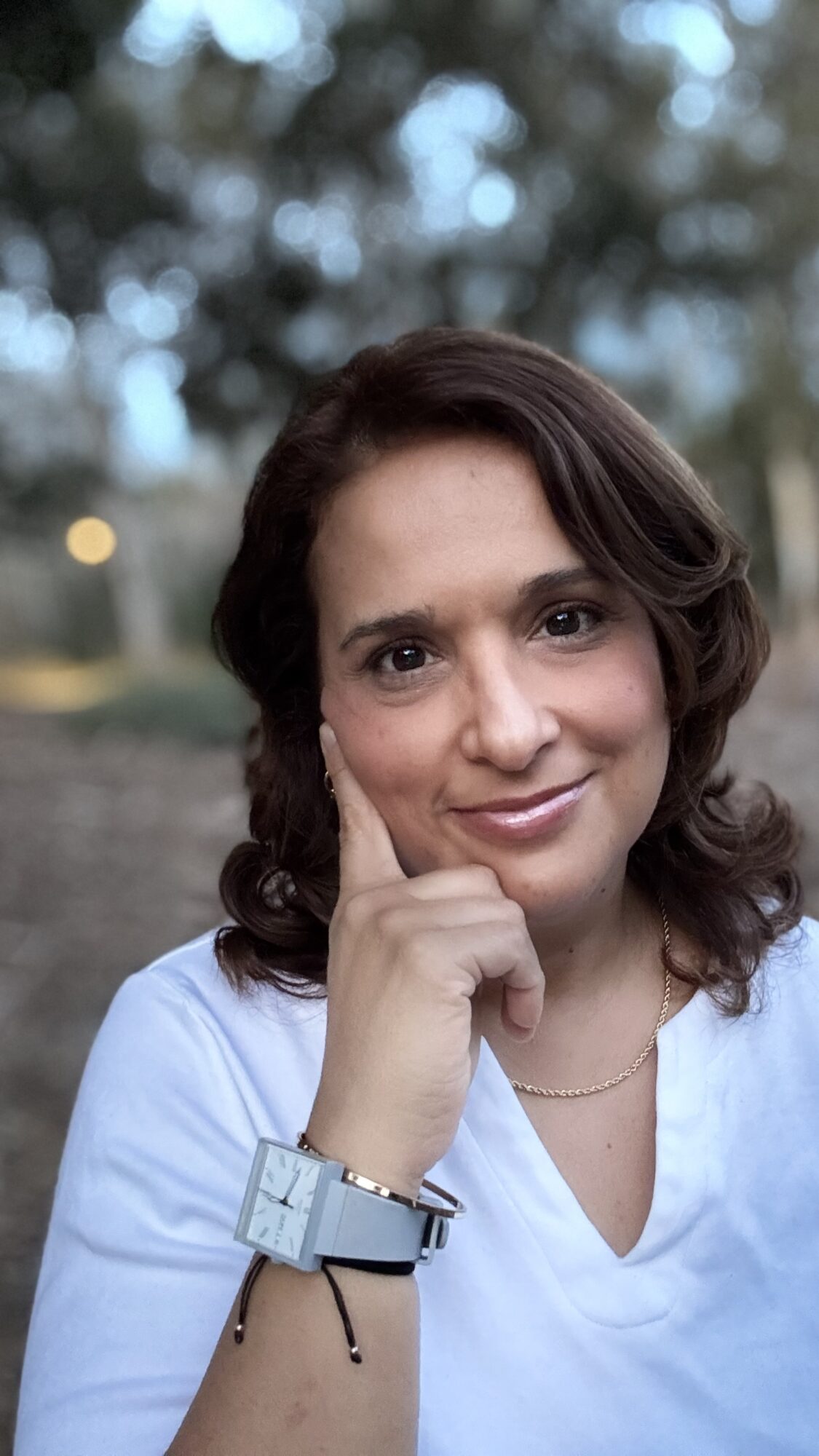 Woman with dark hair smiling outdoors, resting her chin on her hand, wearing a white shirt and watch.