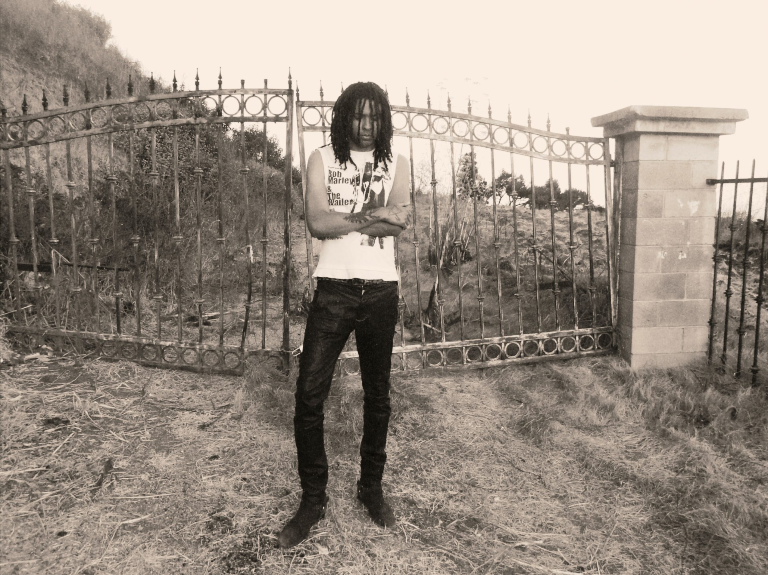 Young man with dreadlocks standing outdoors in front of a metal fence, arms crossed, wearing a sleeveless shirt and jeans.