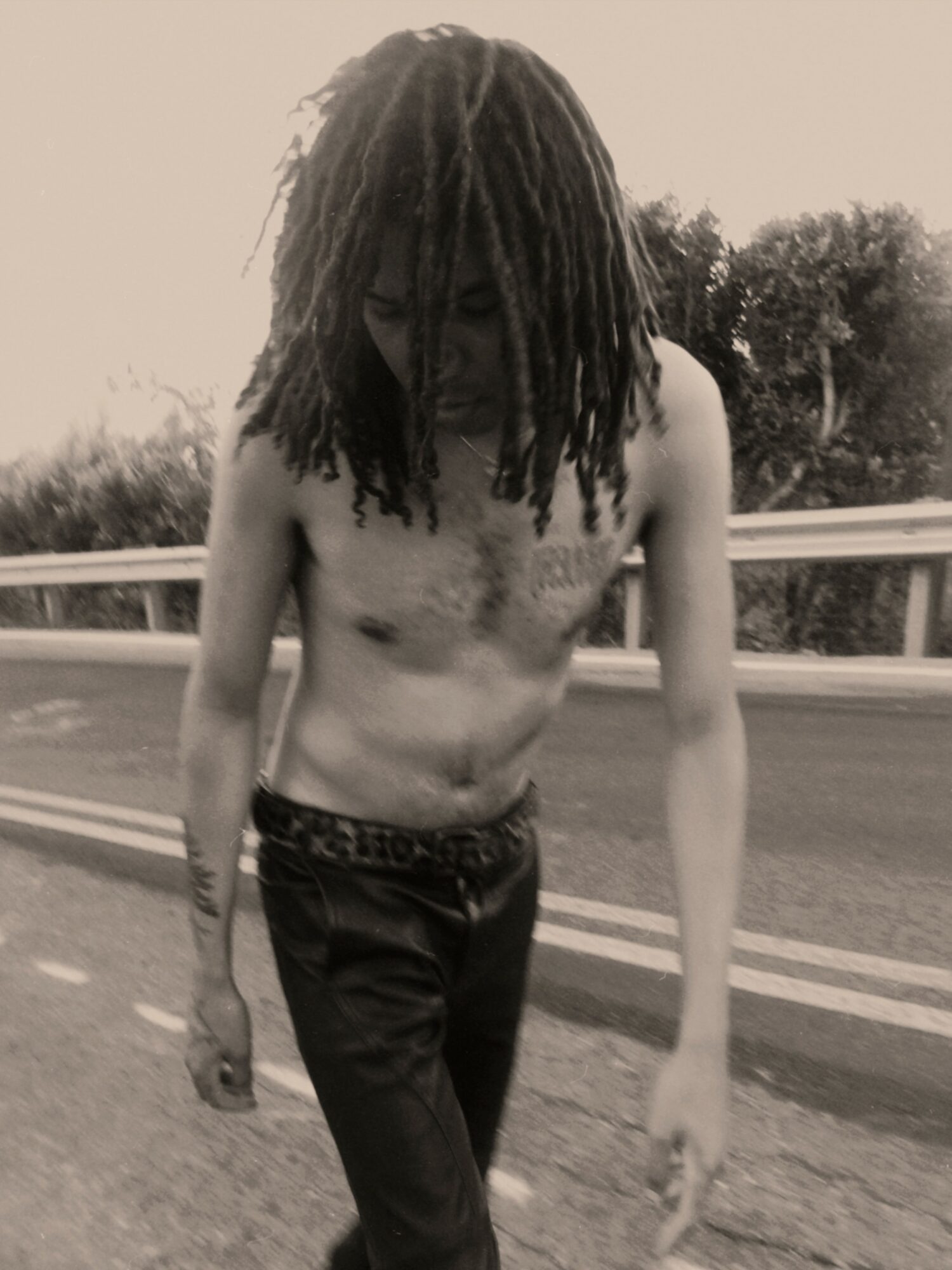Young man with dreadlocks walking shirtless on a road, wearing dark pants, with trees and a guardrail in the background.