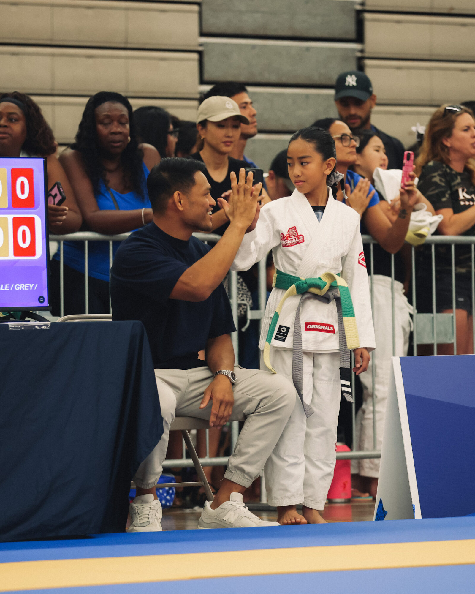 A young girl in a martial arts uniform receives a high-five from a man seated beside her, with spectators in the background.