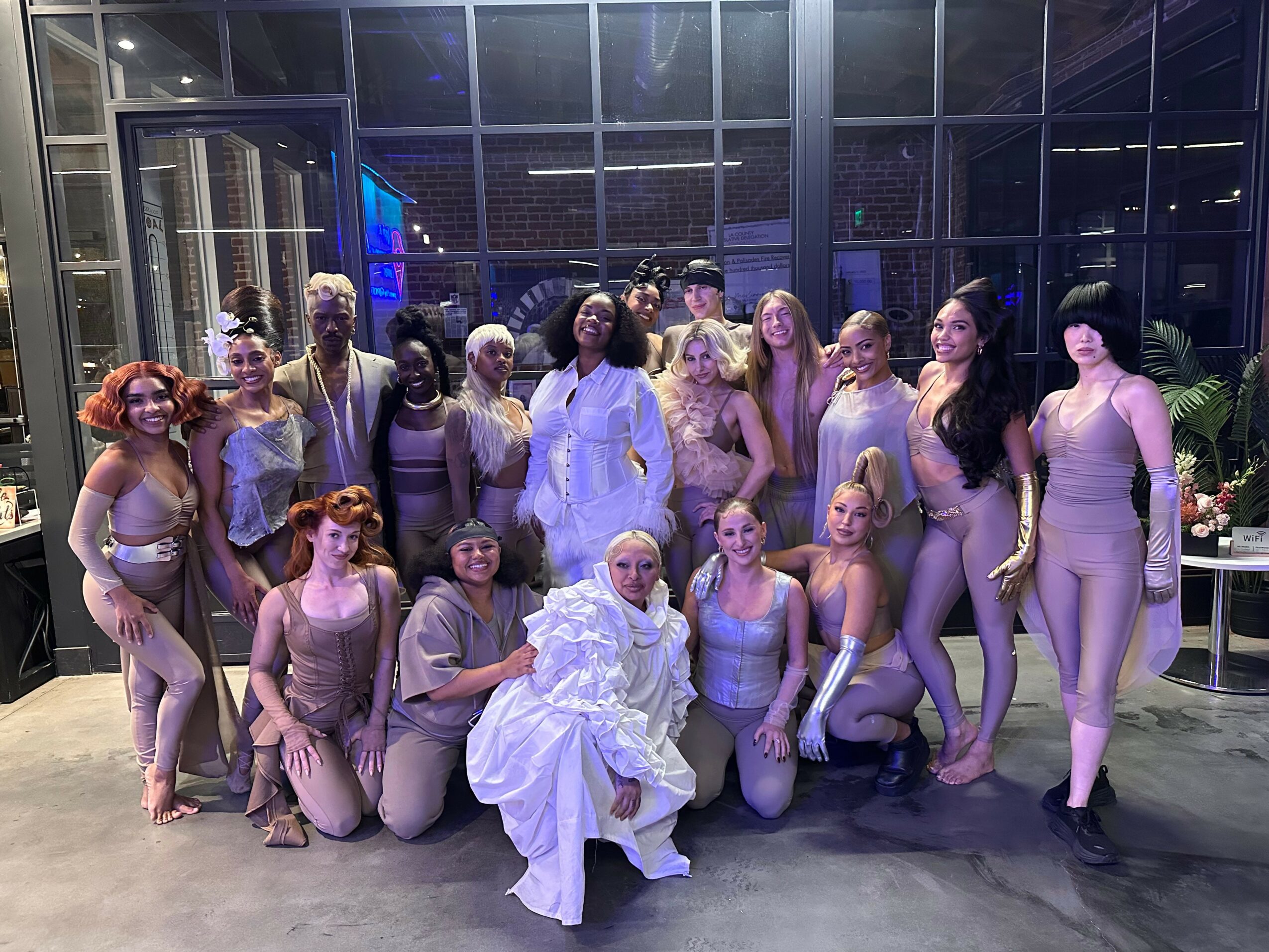 Group of women in costumes and a person in a white dress posing indoors with glass walls and plants.