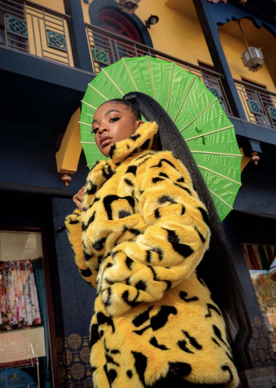 Young woman with long hair holds a green umbrella, wearing a yellow leopard print coat, standing in front of a colorful building.