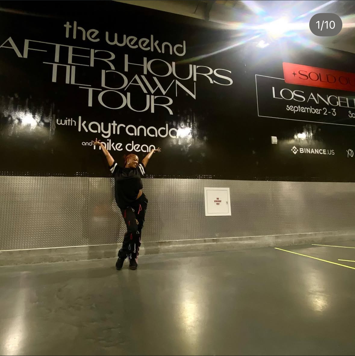 Person standing in front of a large black wall with concert information and a bright light in the top right corner.