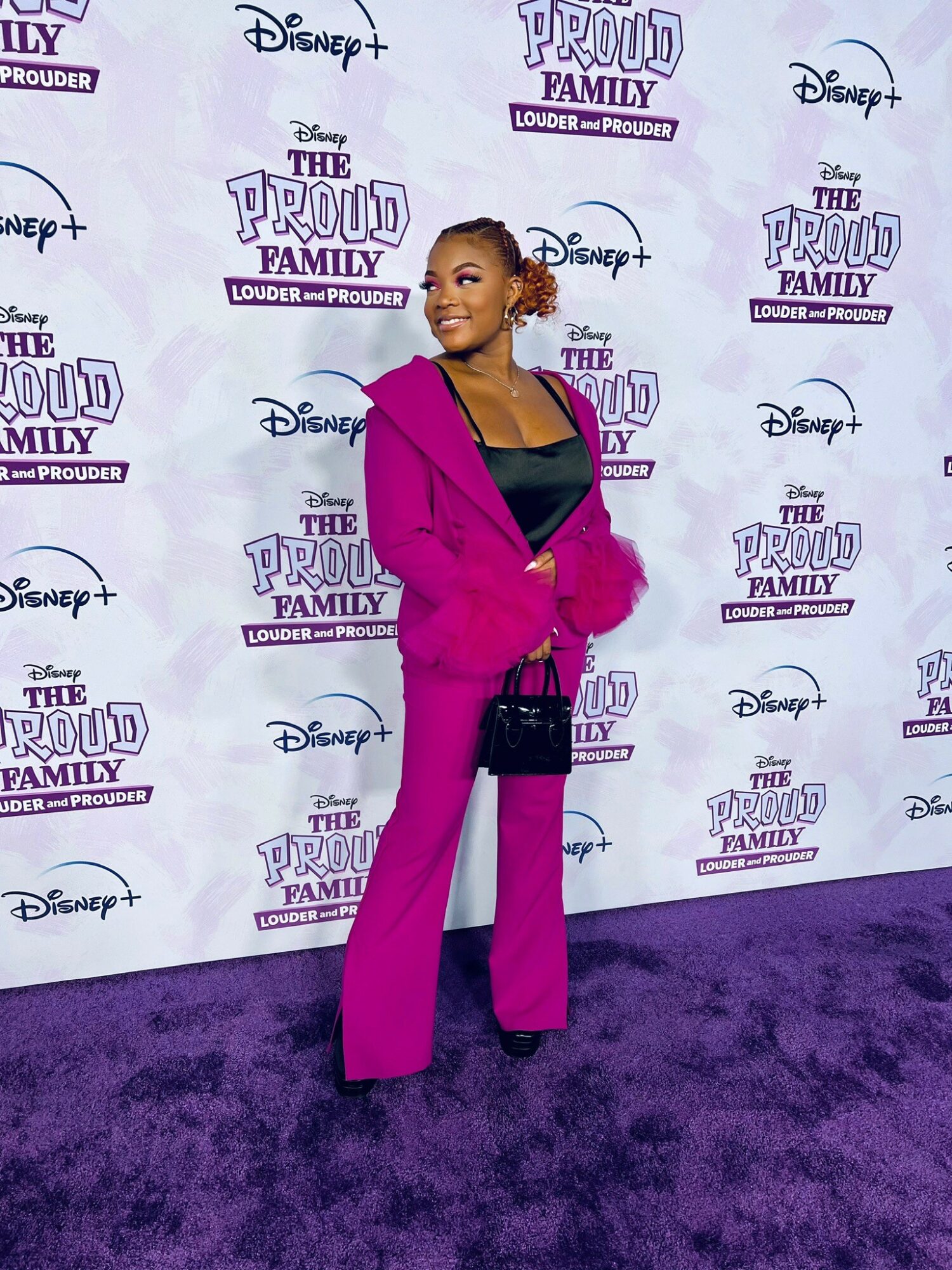 Young woman in pink blazer and white pants standing on purple carpet at Disney event, smiling, with event backdrop behind her.