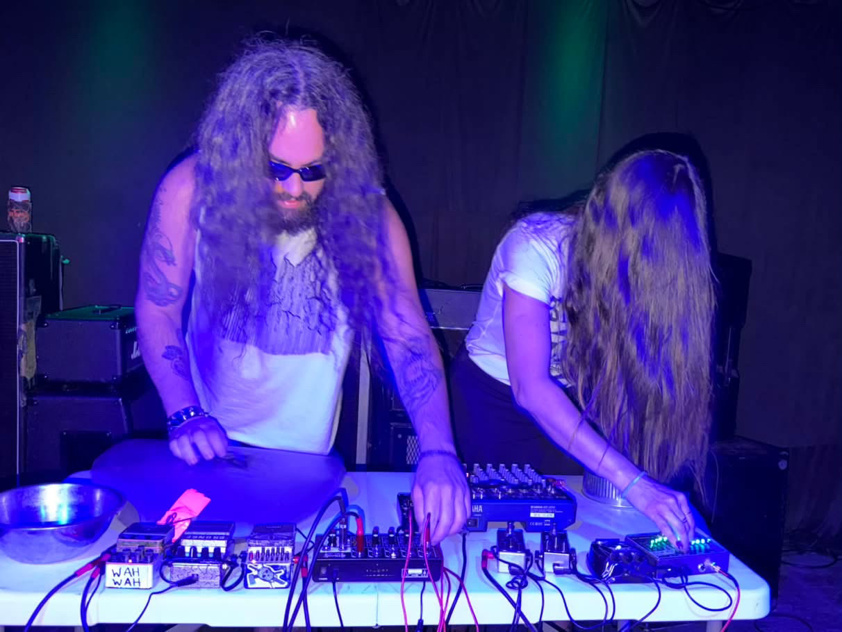 Two people with long hair are working with electronic music equipment on a table, in a dark setting with colorful lighting.