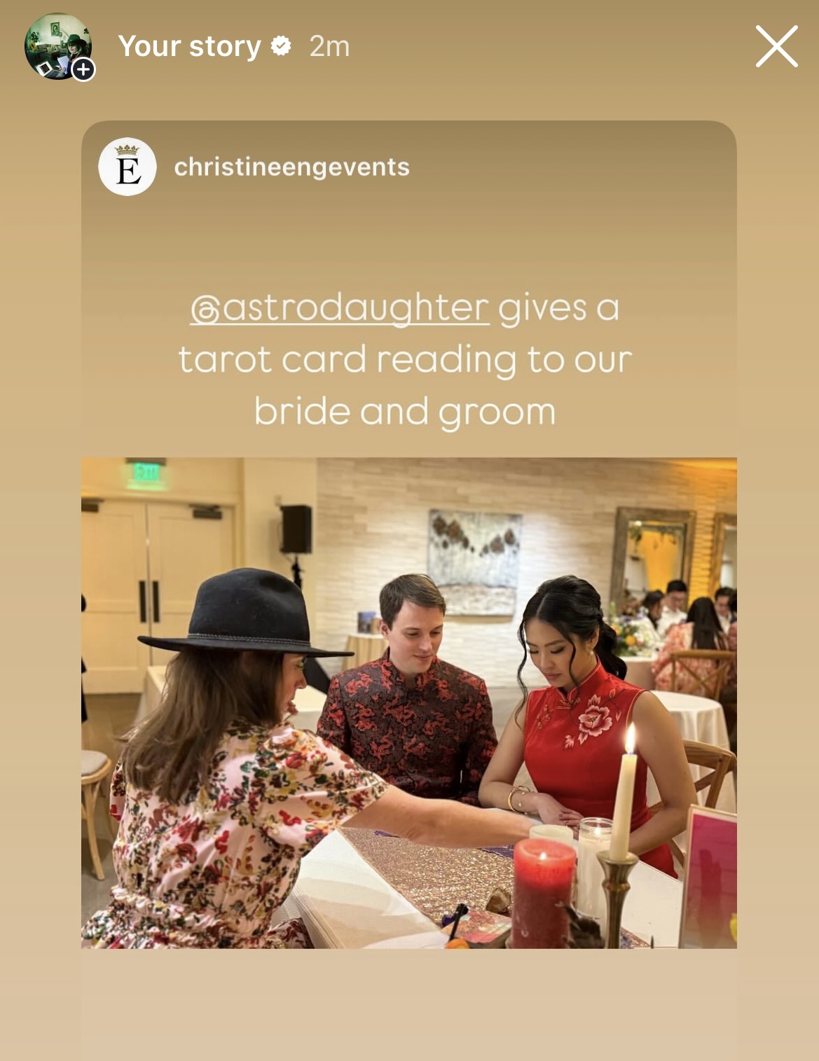 A woman in a floral dress and hat reads a tarot card to a bride and groom at a table in a decorated indoor setting.