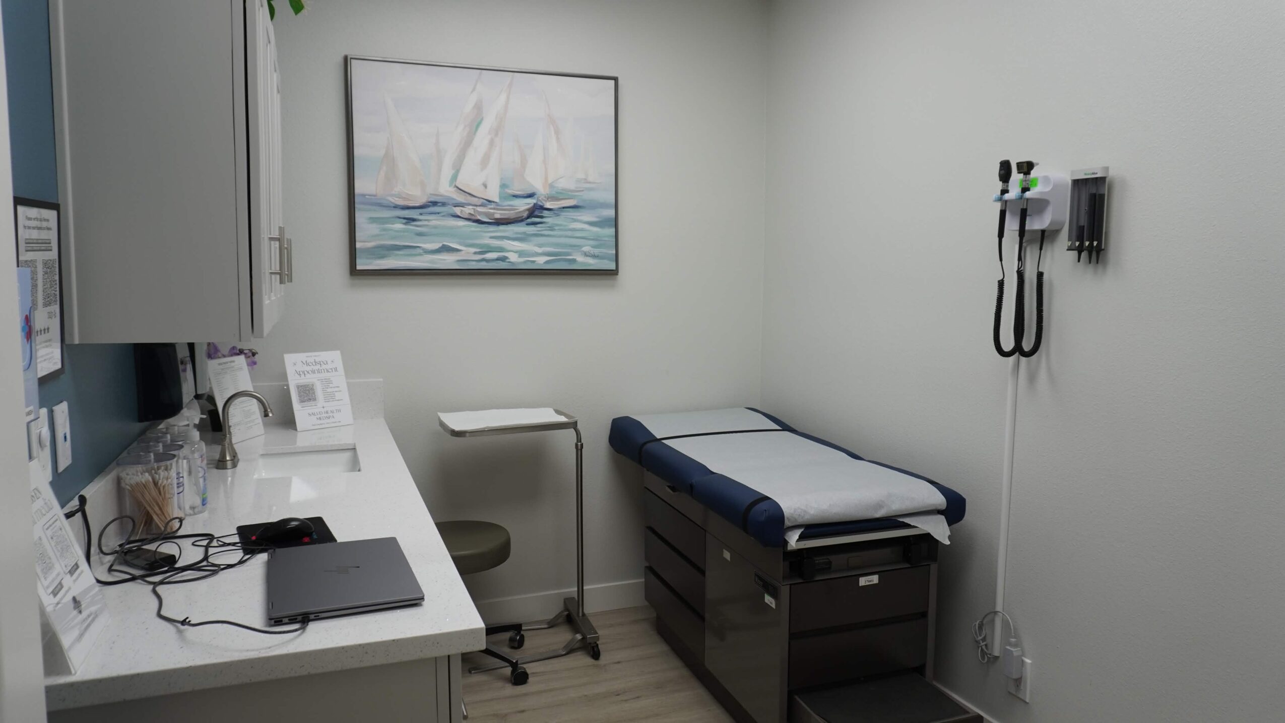 Medical examination room with a bed, desk, and wall-mounted medical equipment, and a painting of sailboats on the wall.