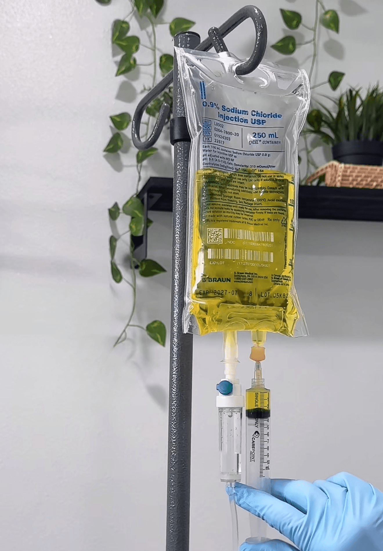 Medical IV bag hanging from a stand with a syringe and tubing, against a white wall with green plants.