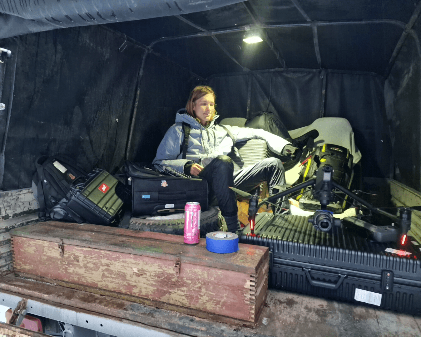 Person sitting in a tent with camera equipment, a motorcycle, and a pink drink can on a wooden box.