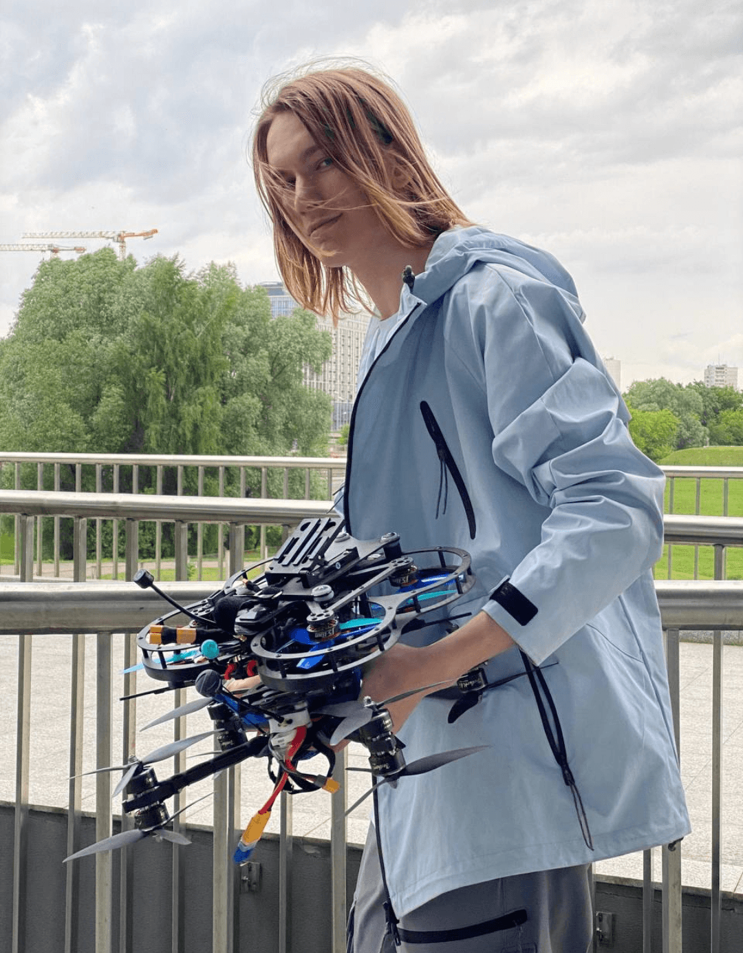 Woman holding a drone outdoors on a bridge with trees and cloudy sky in background.