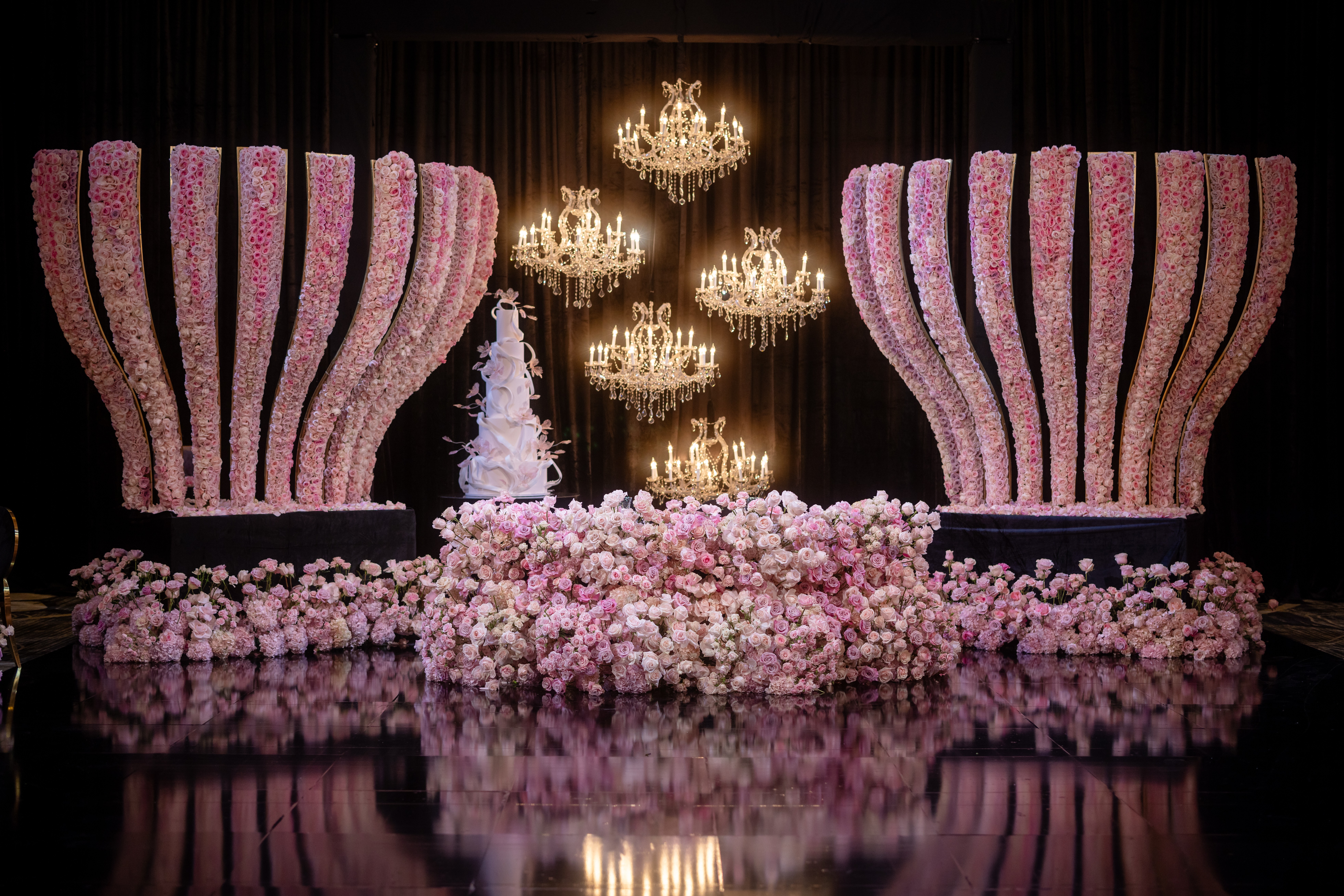 Decorative stage with large pink flower-shaped structures, chandeliers, and a floral arrangement, reflected on a shiny floor.