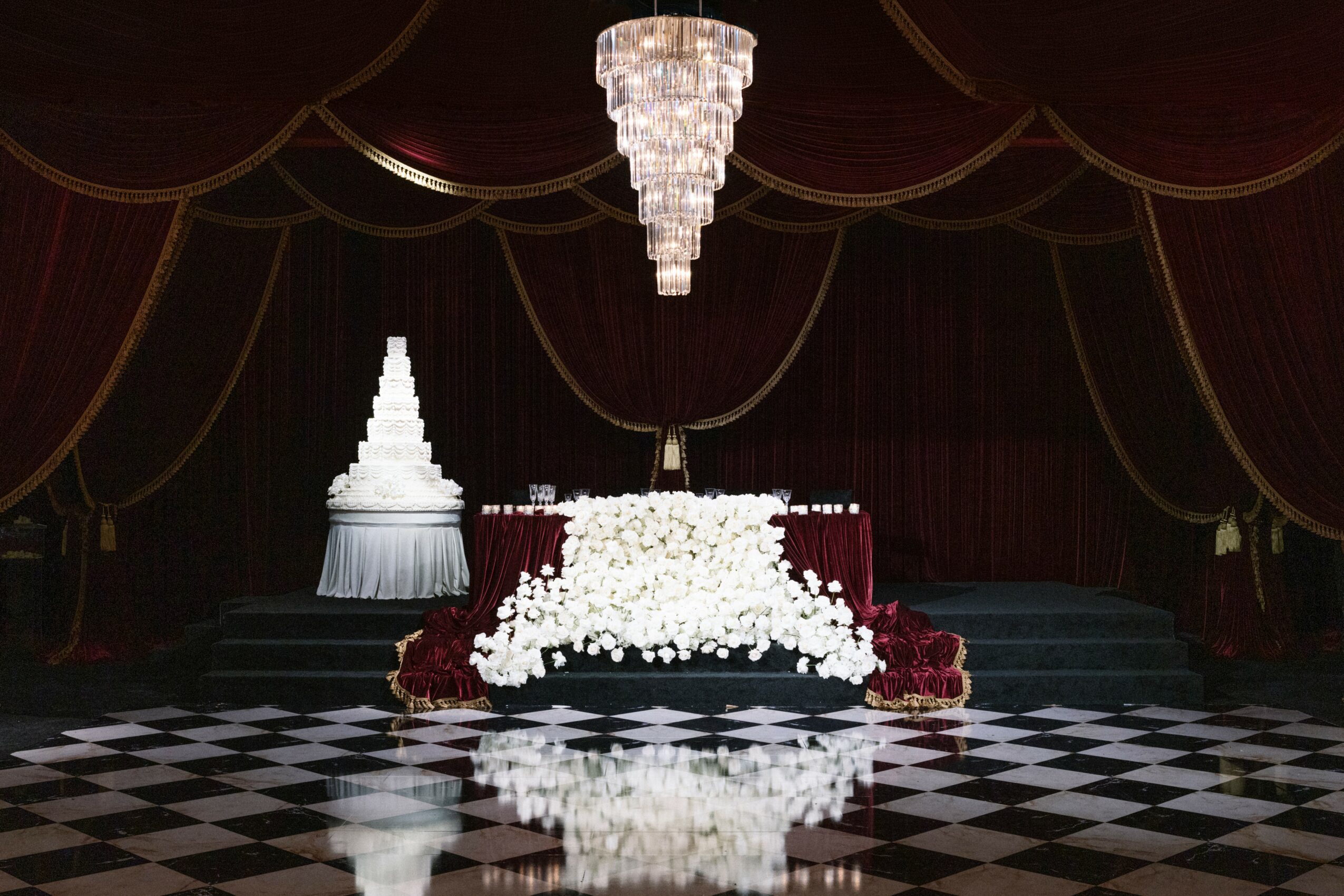 A decorated stage with a large floral arrangement, a cake, and a chandelier in a dark red curtain setting.