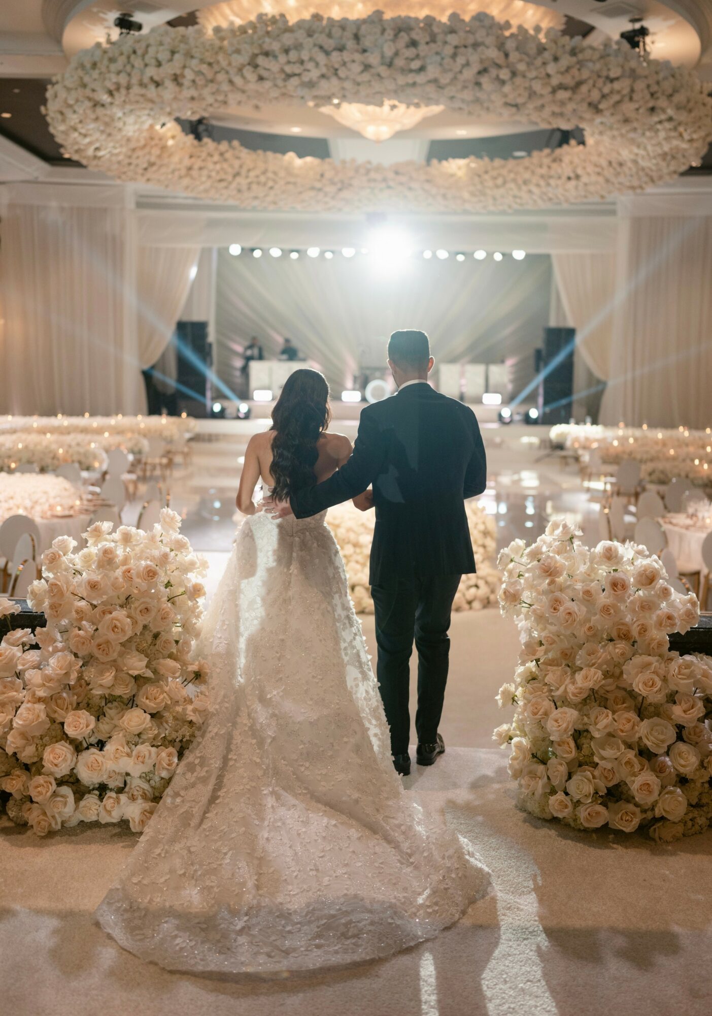 Bride and groom walk down aisle in wedding hall decorated with white flowers and drapery, stage with lights in background.