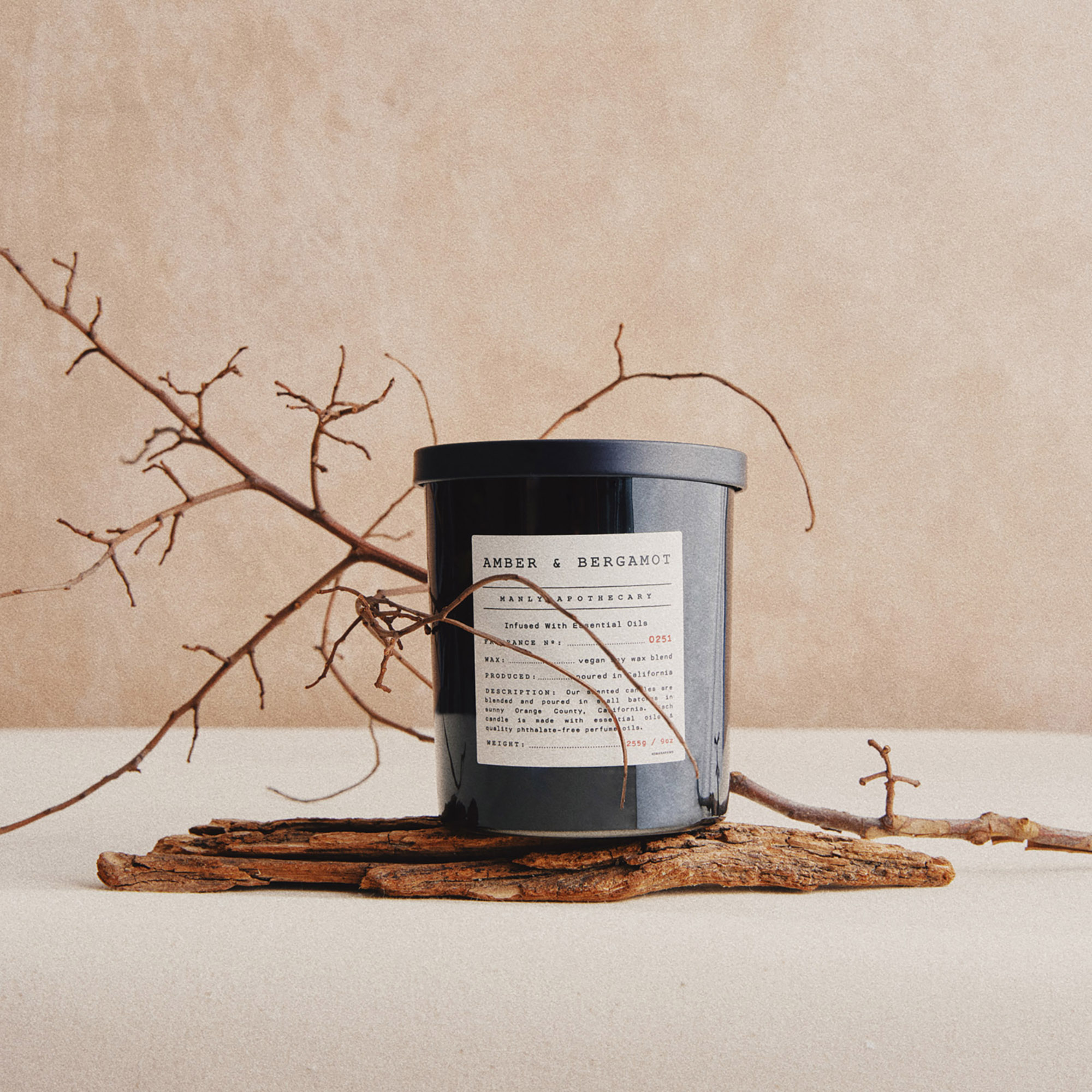 Black candle jar with a label, placed on a piece of wood with a dry branch extending across the background.