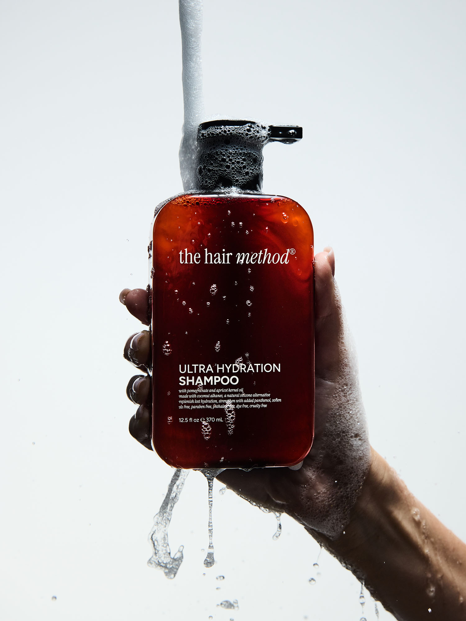 Hand holding a red shampoo bottle with water pouring over it, labeled 'the hair method' and 'ULTRA HYDRATION SHAMPOO'.