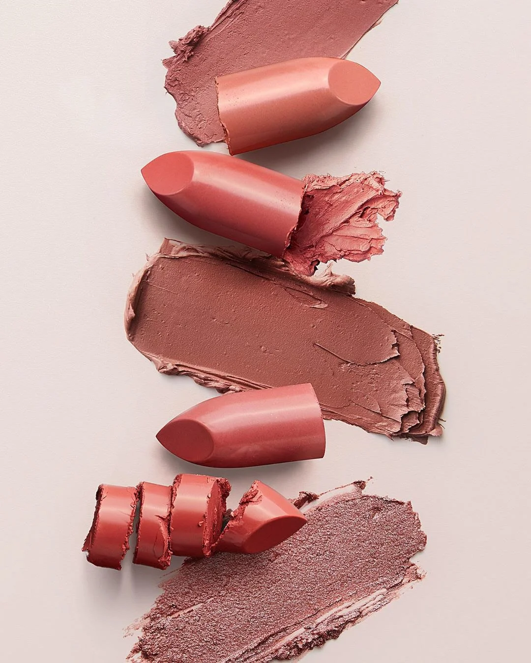 Four lipstick swatches in shades of pink and brown, with a fifth swatch partially broken, on a light background.