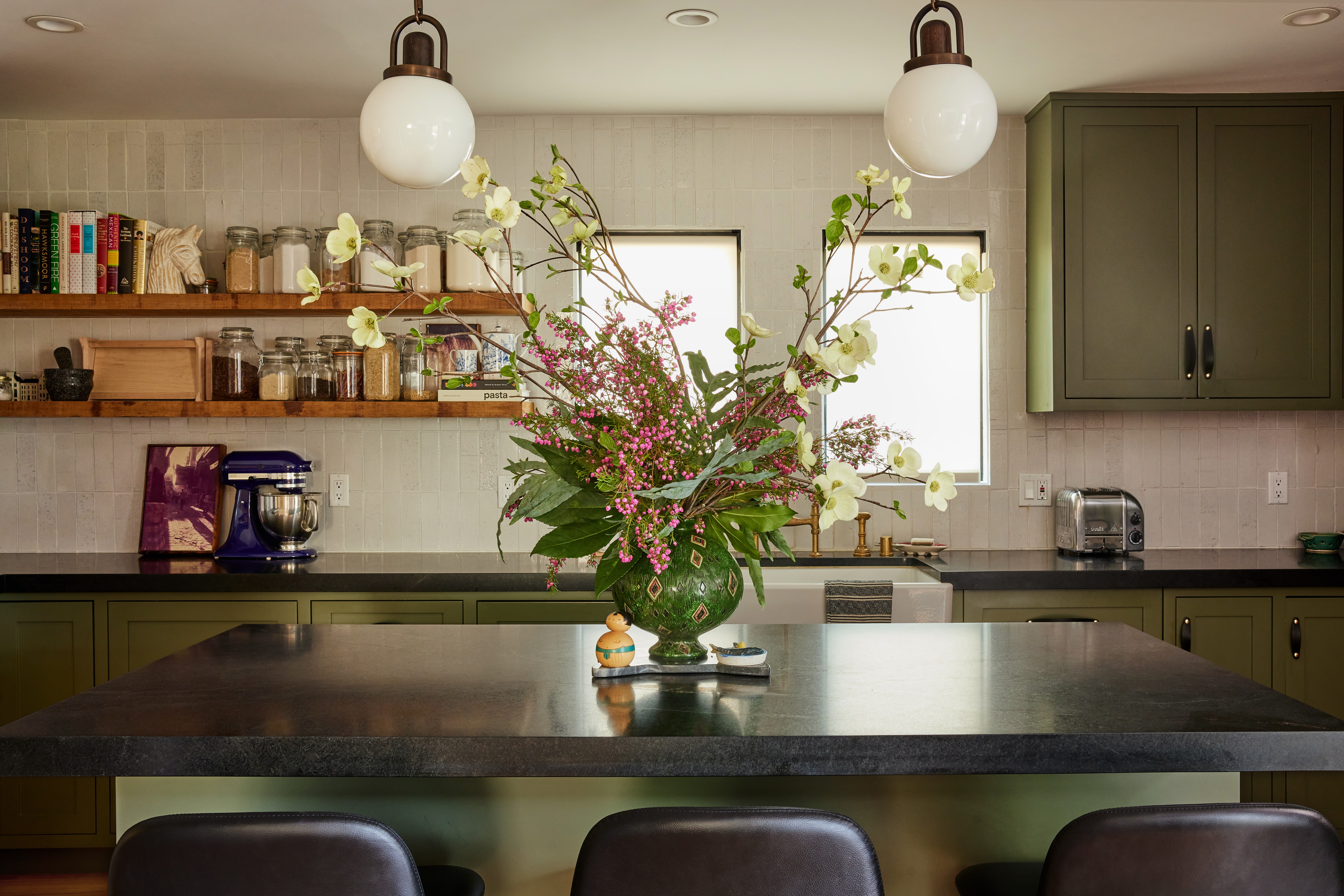 Kitchen with a large flower arrangement on a dark countertop, green cabinets, open shelves, and two hanging lights.