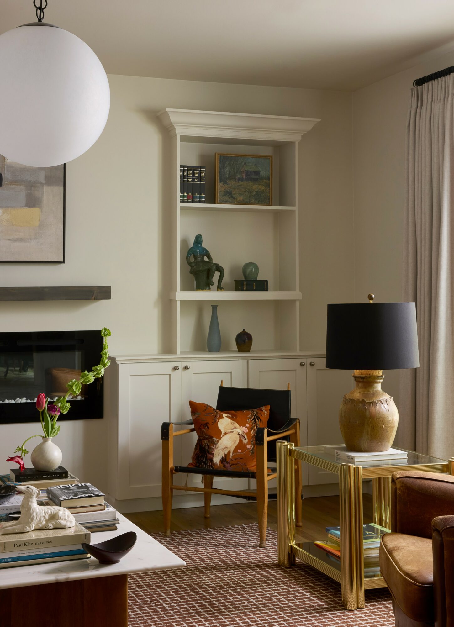 Living room with built-in shelves, a black lamp on a gold table, a wooden chair with a pillow, and a window with curtains.