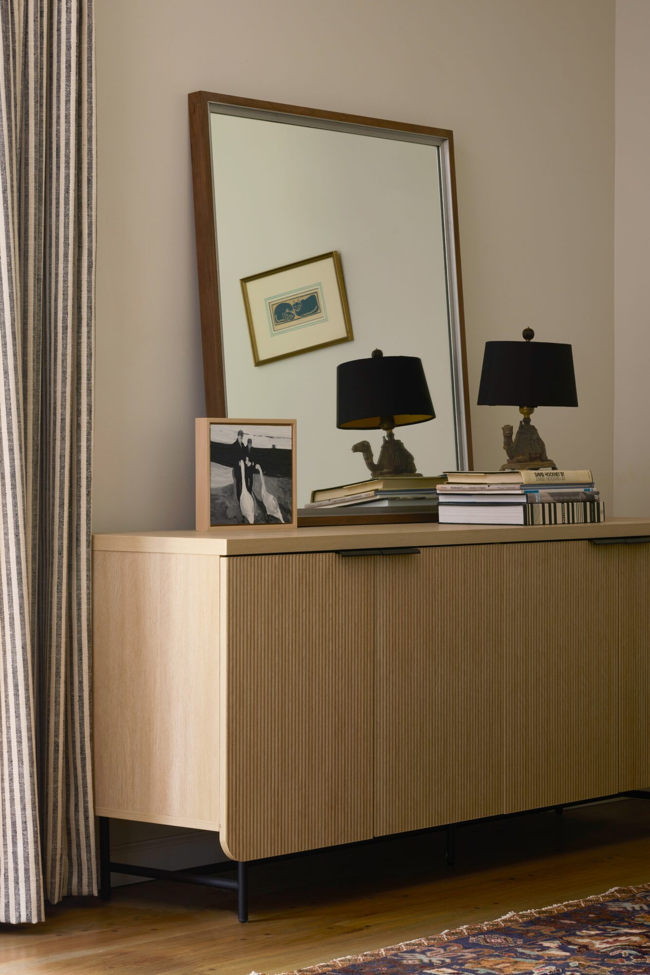 A wooden sideboard with framed pictures, two black lamps, and a mirror reflecting a framed artwork, against a beige wall.